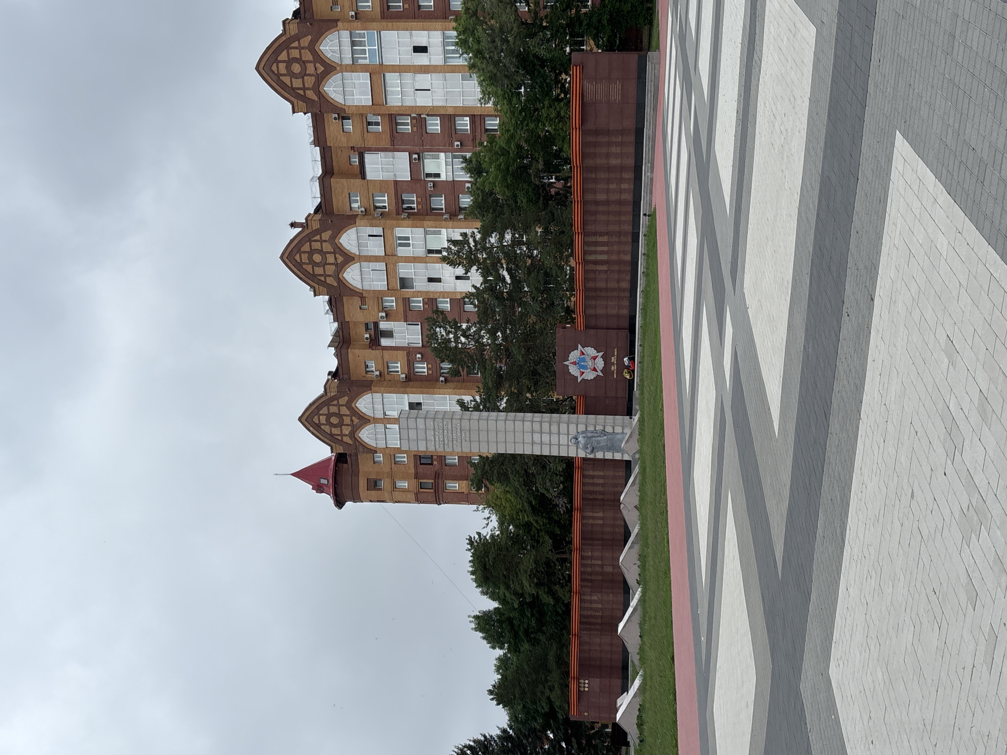 This image depicts a war memorial and a residential building in Blagoveshchensk, Russia. The memorial features a tall, grey obelisk with inscriptions and a statue of a soldier. Behind it, a long, dark red wall is adorned with orange ribbons and a large star emblem. The residential building in the background is made of orange brick with a red-tiled roof and arched windows. The foreground is a paved square with a geometric pattern of grey and white tiles. The sky is overcast.