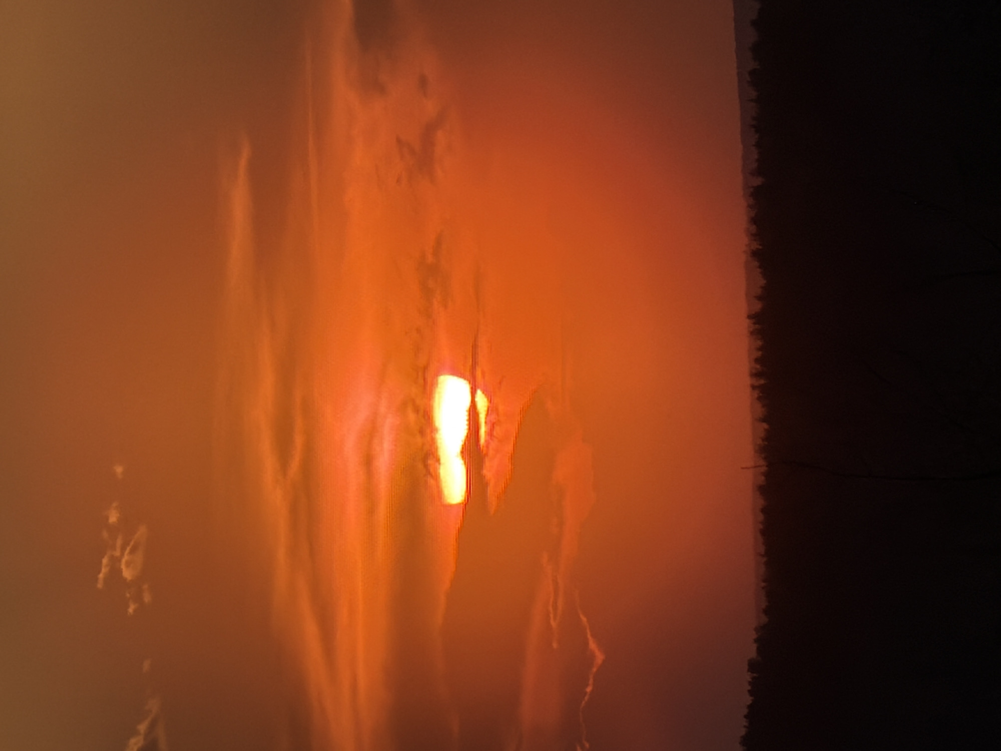 The image depicts a landscape in Chaikovsky, Russia at sunset. The sun is low in the sky, obscured by clouds, resulting in a hazy, orange glow across the scene. To the right, a dark silhouette of a forest line is visible against the orange sky.