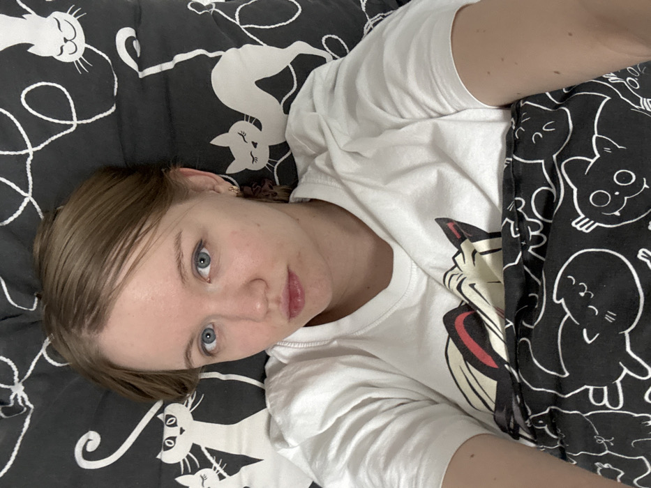 A fair-skinned woman with short, light brown hair and blue eyes lies on a bed. She is angled towards the camera with a neutral expression. She wears a white long-sleeved shirt featuring a graphic design. The bedding includes a black and white comforter with cartoon cats. A second black and white patterned fabric, also featuring cartoon cats, is visible. The overall scene has a relaxed and informal feel.