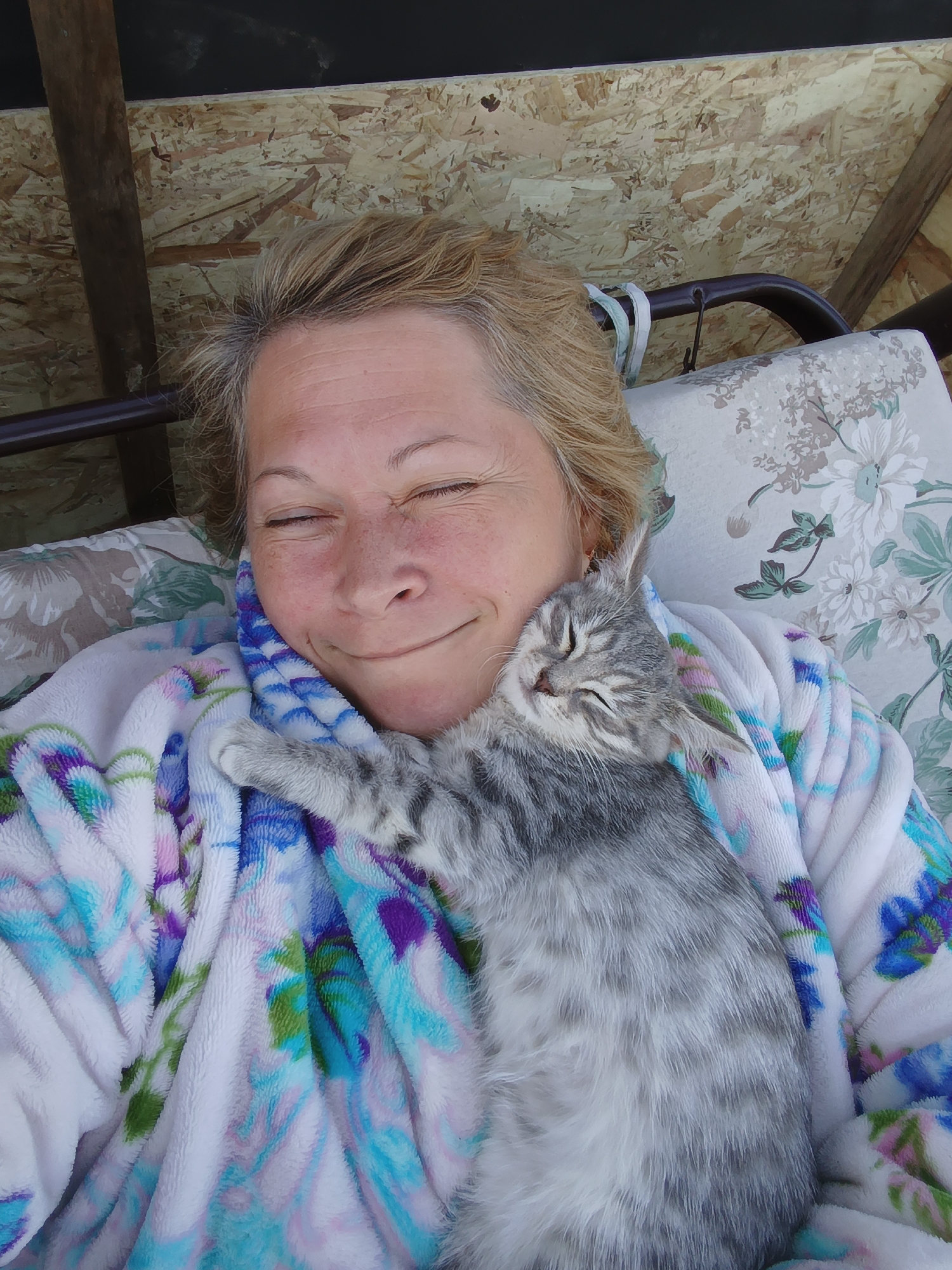 In Ivanovskoe, Russia, a woman is seen resting outdoors, possibly on a swing seat, with her eyes closed and a contented expression. She is wearing a fluffy bathrobe with a floral pattern. Cuddled closely against her neck is a small, gray tabby kitten, also with its eyes closed, appearing to be asleep. The background suggests an outdoor setting with wooden paneling and a glimpse of a floral-patterned cushion on a swing. The overall scene conveys a sense of peace and affection between the woman and the kitten.