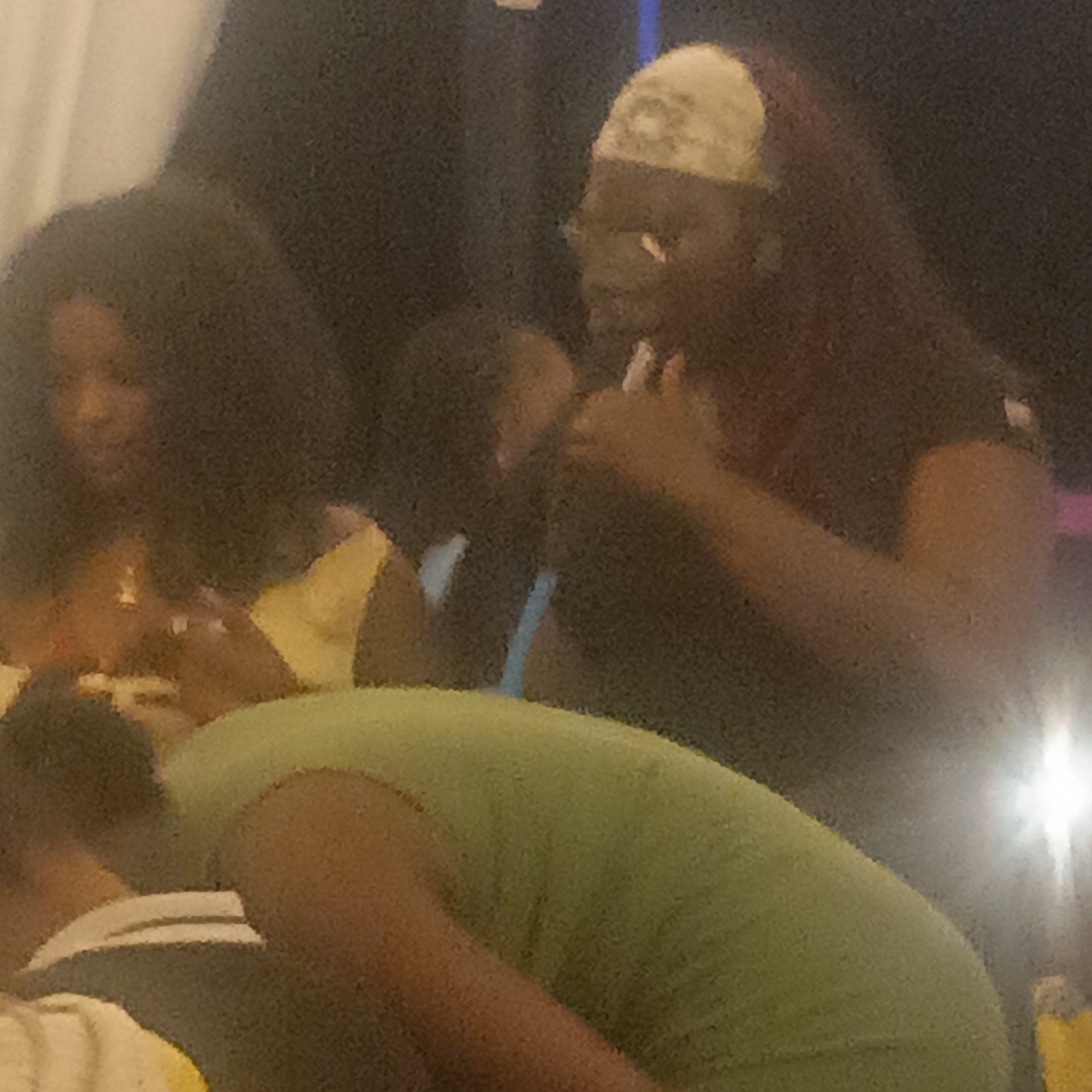 In a dimly lit setting, possibly Ube, Nigeria, several individuals are gathered. A woman with dark, curly hair is partially visible, wearing a yellow top. Another woman with reddish-brown hair and a head covering is observed holding a cigarette to her mouth. A person in a green shirt is in the foreground, partially obscuring the view. The environment appears to be indoors with a light source creating glare in the background.