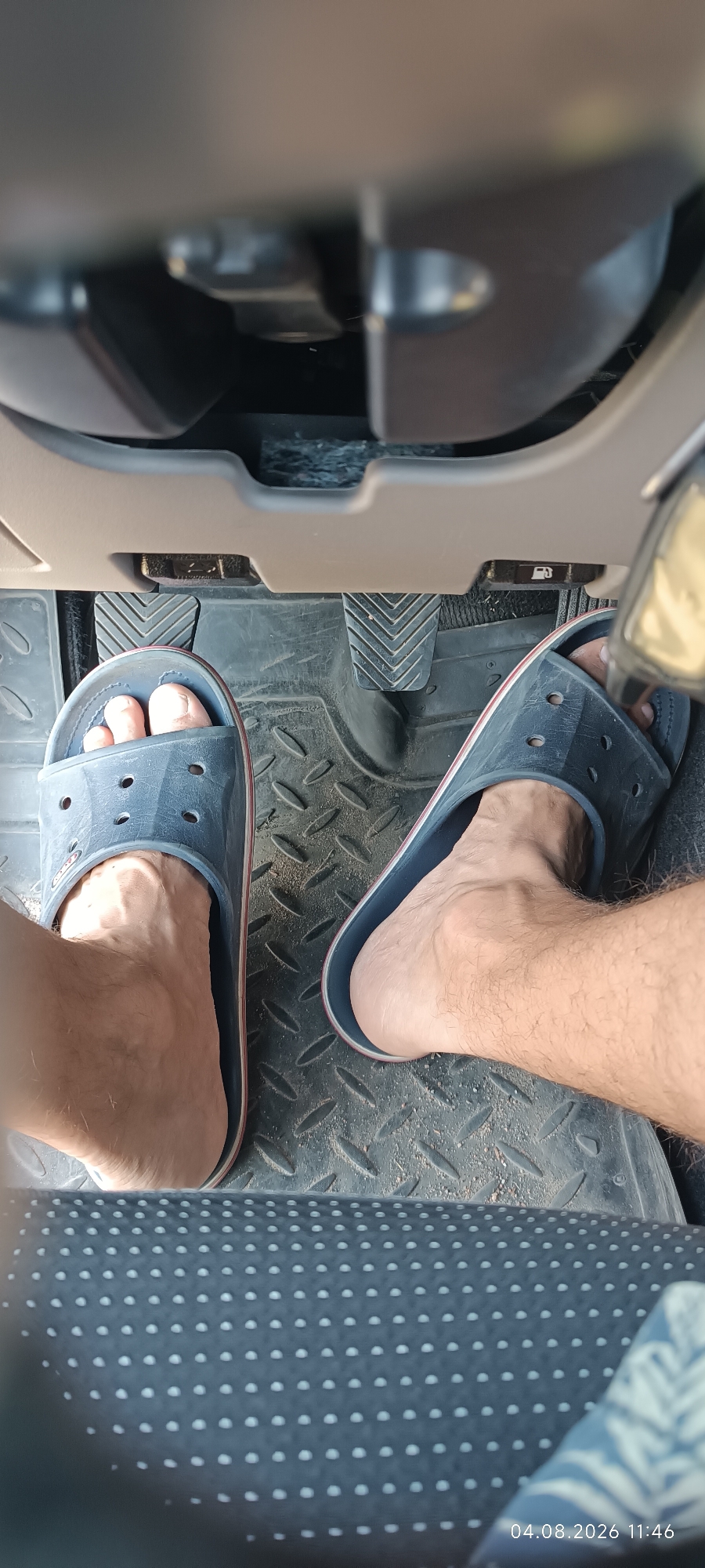 The image shows a close-up view of a person's feet wearing blue flip-flops inside a vehicle. The feet are positioned over the car's pedals, which are visible on a black rubber mat. The interior of the car is dimly lit. The image was taken on August 4, 2026, at 11:46 AM, in Starokozache, Ukraine.