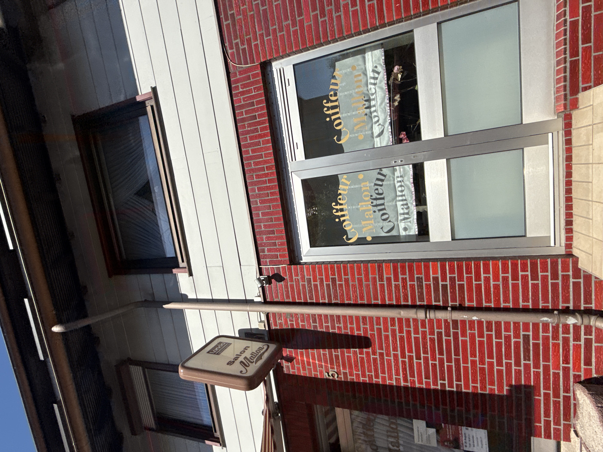 This image captures a storefront in Bad Sachsa, Germany, identified by the sign "Salon Mallon Coiffeur". The building features red brickwork on the lower level and a light-colored facade on the upper level. A tall, gray downspout runs vertically along the brick wall. The storefront windows display text that reads "Coiffeur Mallon Coiffeur", indicating a hair salon. Above the storefront, there are two windows on the upper floor, one with blinds pulled down. The sky is clear and blue, suggesting a sunny day. The overall scene presents a typical urban commercial building.