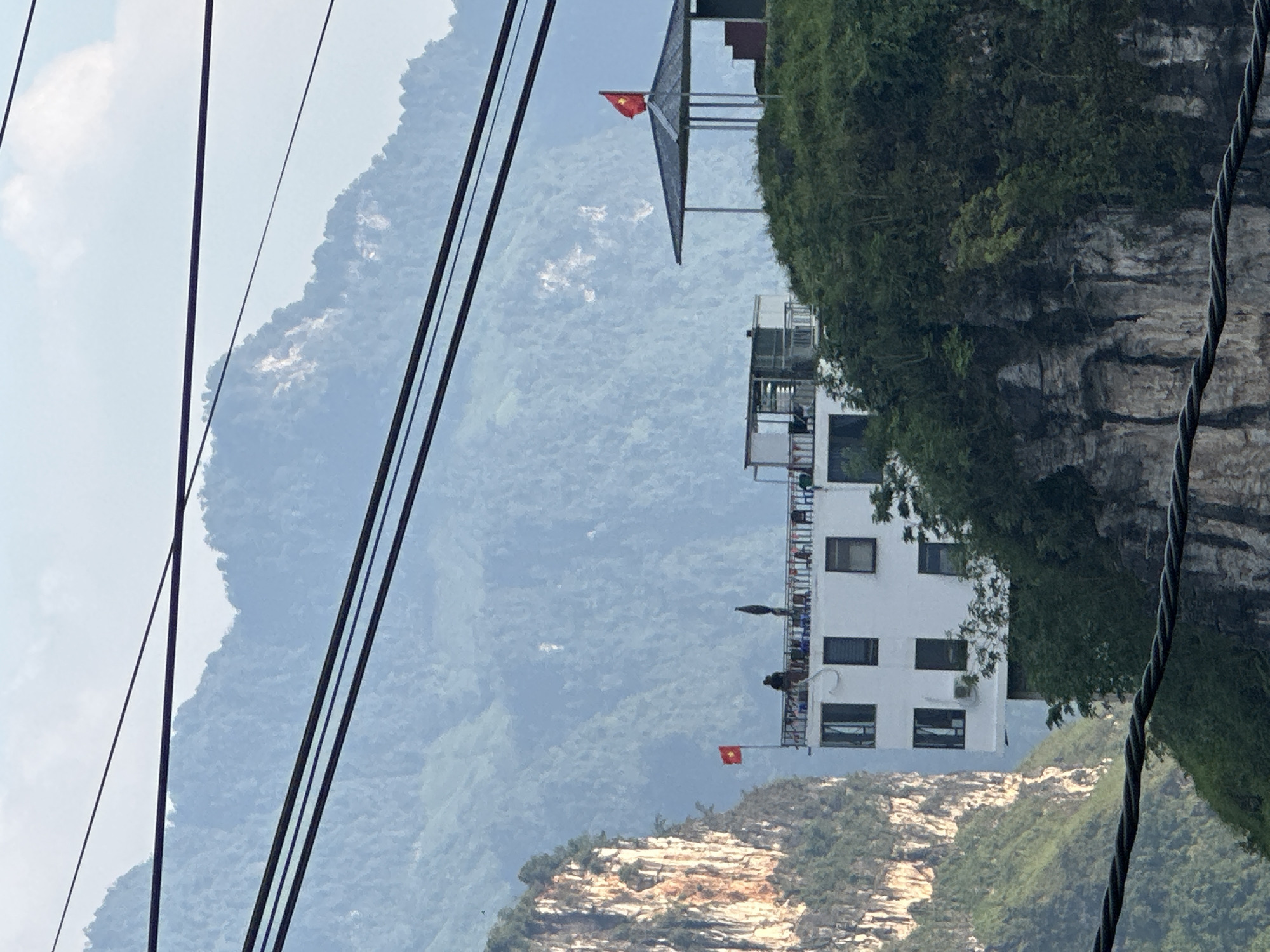 The image shows a multi-story building perched on a cliffside in Đường Thượng Commune, Vietnam. A Vietnamese flag is visible at both the top and bottom of the building. The building is white with dark windows, and the cliffside is covered in vegetation. The background features a mountainous landscape with cable lines running across the frame.