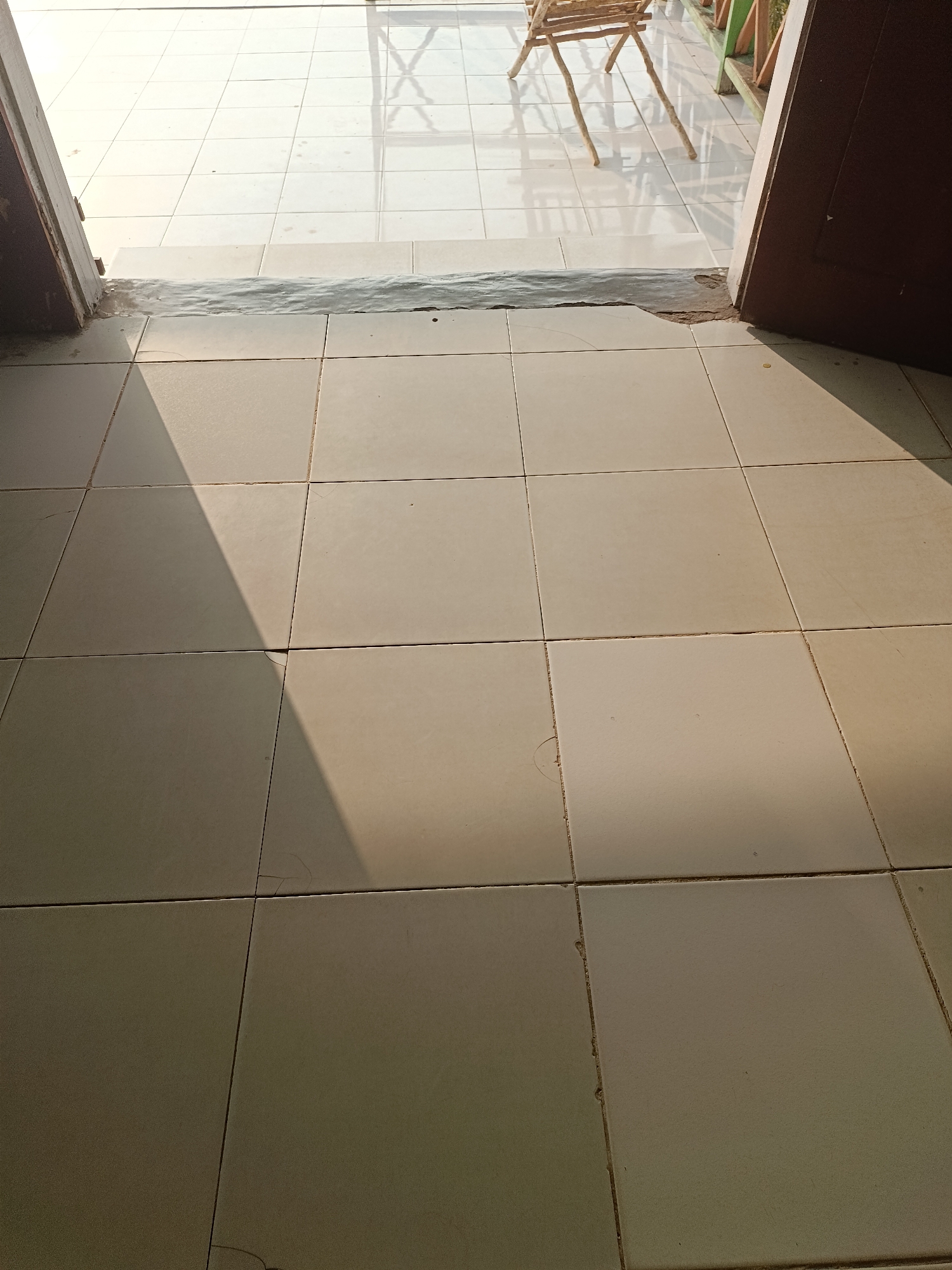 The image shows a tiled floor in what appears to be an outdoor area, possibly a patio or veranda, with sunlight casting shadows. The tiles are square and light-colored. In the background, a wooden structure resembling a chair or table can be seen, along with some railing. The scene depicts an entryway, with a dark-colored door frame visible on the right and another on the left, partially open. The location is identified as Mansalean, Indonesia. The overall impression is a snapshot of a typical outdoor space in a residential or commercial setting, bathed in sunlight.