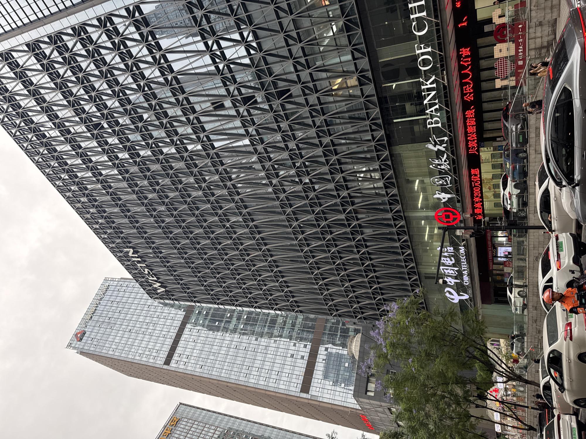 The image depicts a street scene in Yuzhong District, Chongqing, China, dominated by tall, modern buildings and busy traffic. The Westin Chongqing Liberation Square hotel is visible, recognizable by its geometric facade. The Bank of China and China Telecom are also identified through their signage. Several white cars are lined up on the street, and a delivery driver wearing an orange jacket is visible. A tree with light purple flowers sits near the China Telecom signage. The overcast sky contributes to the scene's muted lighting.