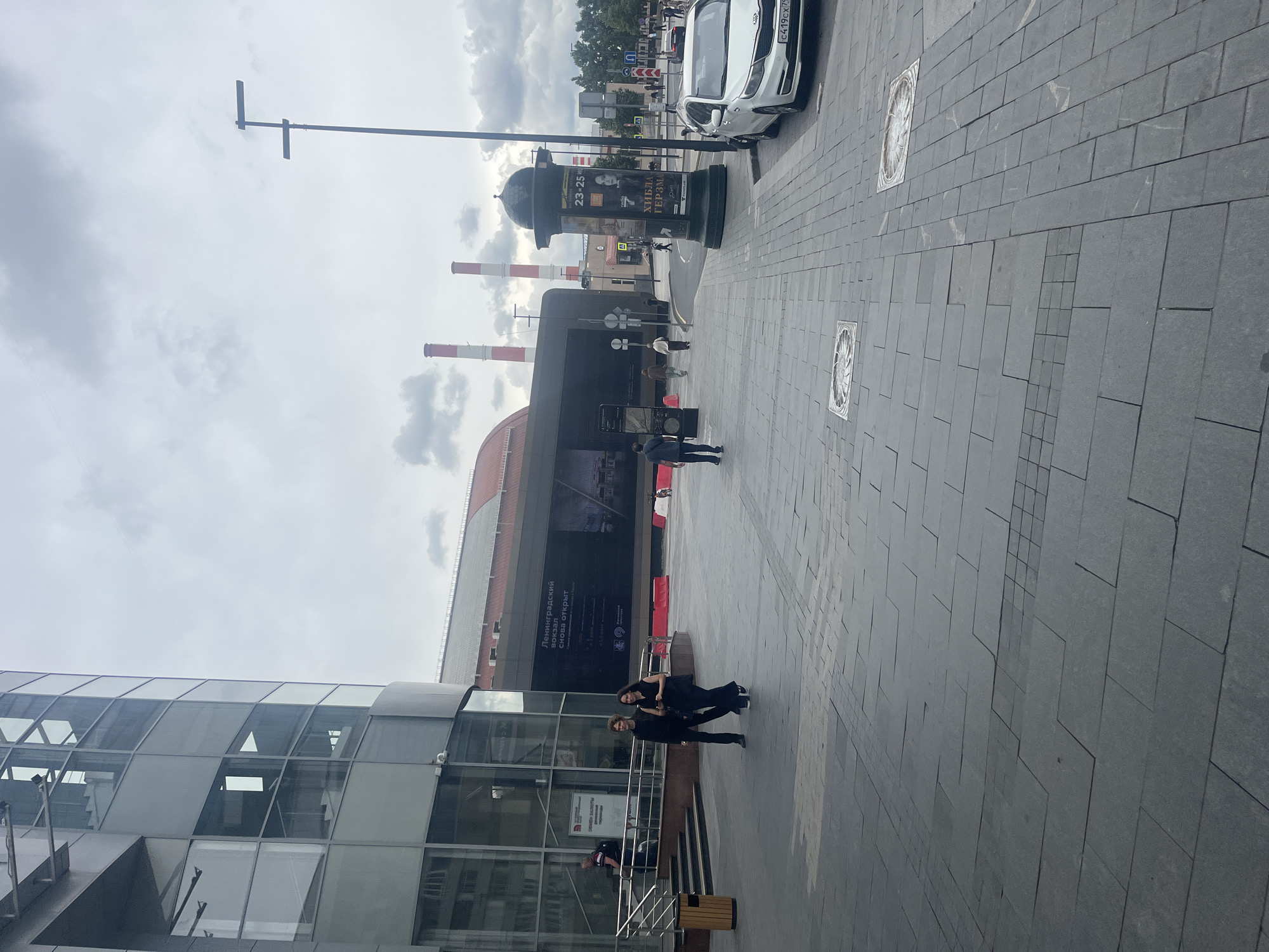This is a medium-shot, eye-level photograph of an outdoor urban scene in Moscow, Russia, likely at Leningradsky Station. In the foreground, a paved plaza with a gray, patterned stone surface dominates the lower half of the frame. Two square manhole covers with ornate designs are visible on the plaza. To the left, a modern glass-and-metal building features a curved facade with multiple windows and a recessed entrance with a set of stairs leading up. A dark metal railing lines the stairs, and a trash receptacle is situated at the bottom. A person in a dark shirt and backpack walks up the stairs.
Further into the midground, two individuals, a man with curly hair wearing a dark jacket and trousers and a woman with dark hair wearing a dark dress, walk towards the right of the frame. Behind them, a large, dark billboard with Russian text is displayed on the side of a long, reddish-brown building that appears to be part of the station complex. Two tall, industrial-looking smokestacks with red bands on their upper sections rise into the overcast sky. A street lamp with two horizontal lights stands to the right of the smokestacks. A dark green, cylindrical kiosk with posters, including one featuring a woman's face and text in Russian, is prominently placed on the plaza.
In the background, a white car is parked on the right side of the street, with a license plate visible. The street continues to the right, lined with trees and more buildings. The sky is heavily overcast with light gray clouds. The overall impression is a slightly dreary, cloudy day in a bustling urban environment.