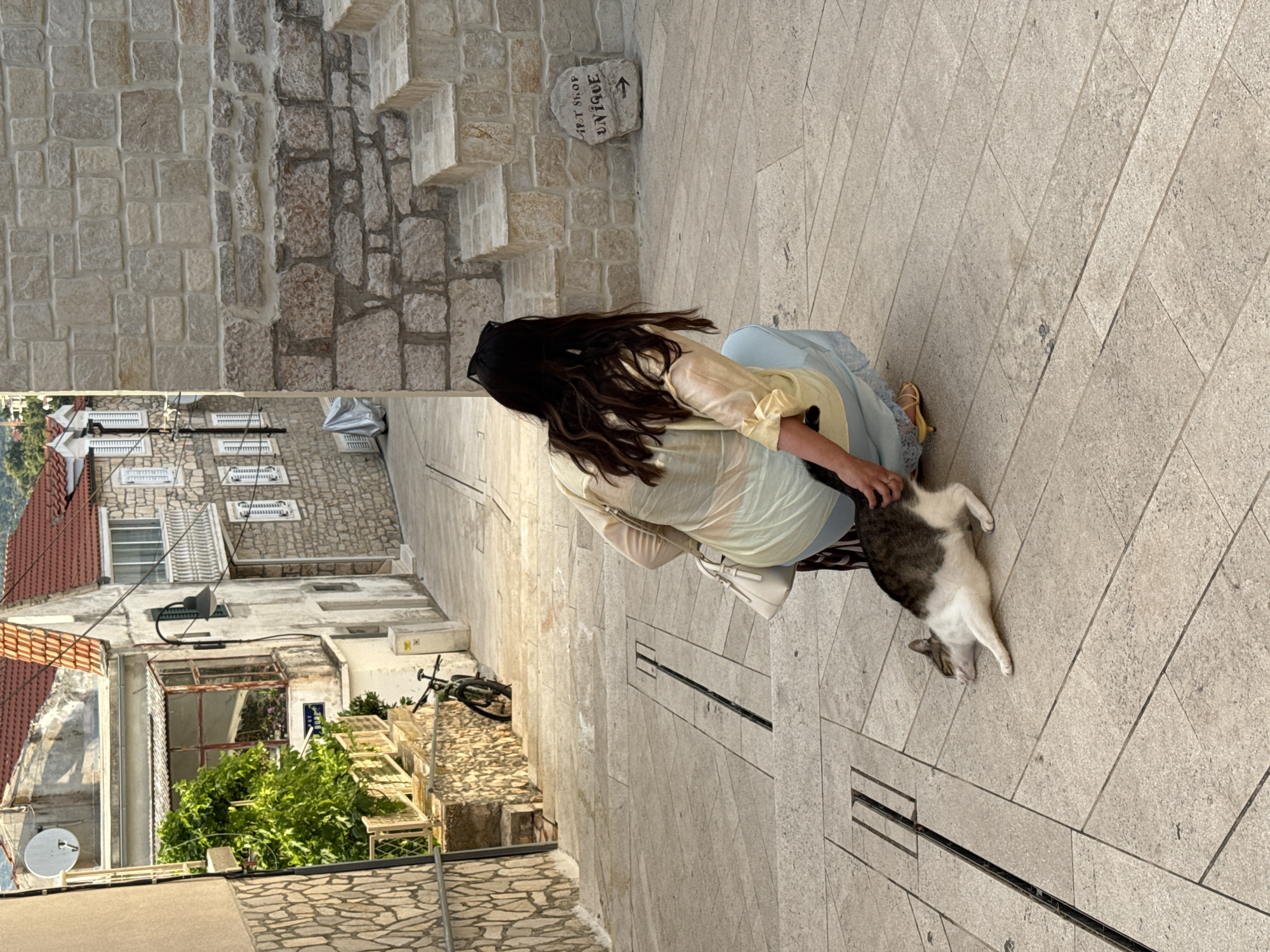 A woman wearing a sheer yellow top and a light blue skirt crouches down to pet a tabby and white cat lying on a cobblestone street in Primošten, Croatia. The cat is stretched out on its side with its eyes closed, appearing relaxed. The woman has long, dark hair and is wearing sunglasses on her head. She is holding a white purse with a gold strap. Behind the woman, a stone wall with steps leads up to a building. A sign on a rock by the steps reads "SPOT SHOP UNIQUE" with an arrow pointing right. In the background, a narrow street winds uphill, lined with stone buildings with red tile roofs. A bicycle is leaning against a wall on the left side of the street. The overall scene depicts a quiet moment in a European coastal town.