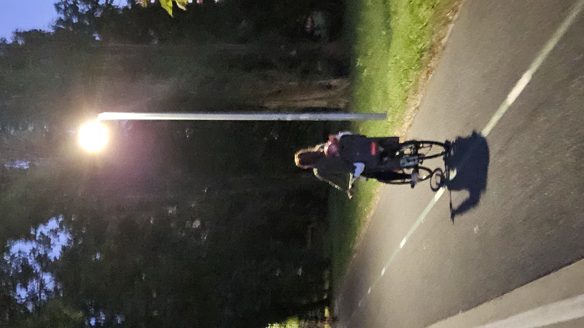 A person is riding a bicycle on a paved path at dusk, illuminated by a streetlamp. The person is wearing a dark jacket and has long brown hair. A child seat is attached to the back of the bicycle, and a dark bag is in the seat. The path is lined with green grass and tall trees. The streetlamp is a tall, white pole with a bright, circular light at the top. The scene is cast in a soft, evening light, creating shadows on the path. The location is identified as being in the city of Prešov, Slovakia.