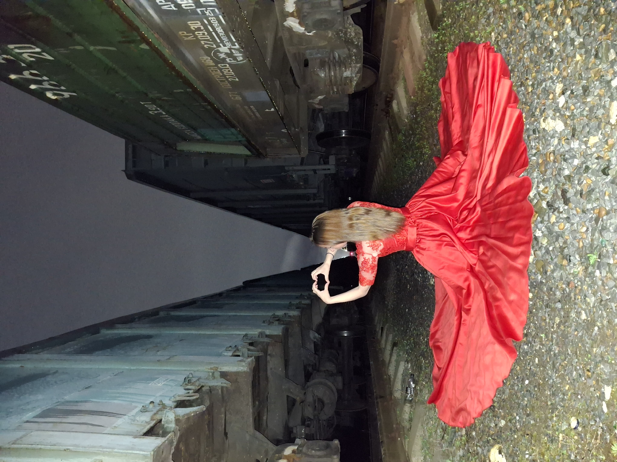 A woman in a red dress stands between two train cars in an undisclosed location in the city of Unknown, Unknown, appearing to take a photo with her phone. The train cars are weathered, with exposed metal and faded markings. The ground is covered in gravel and sparse vegetation. A plastic water bottle sits near the woman's dress. The sky above the train cars is dark.