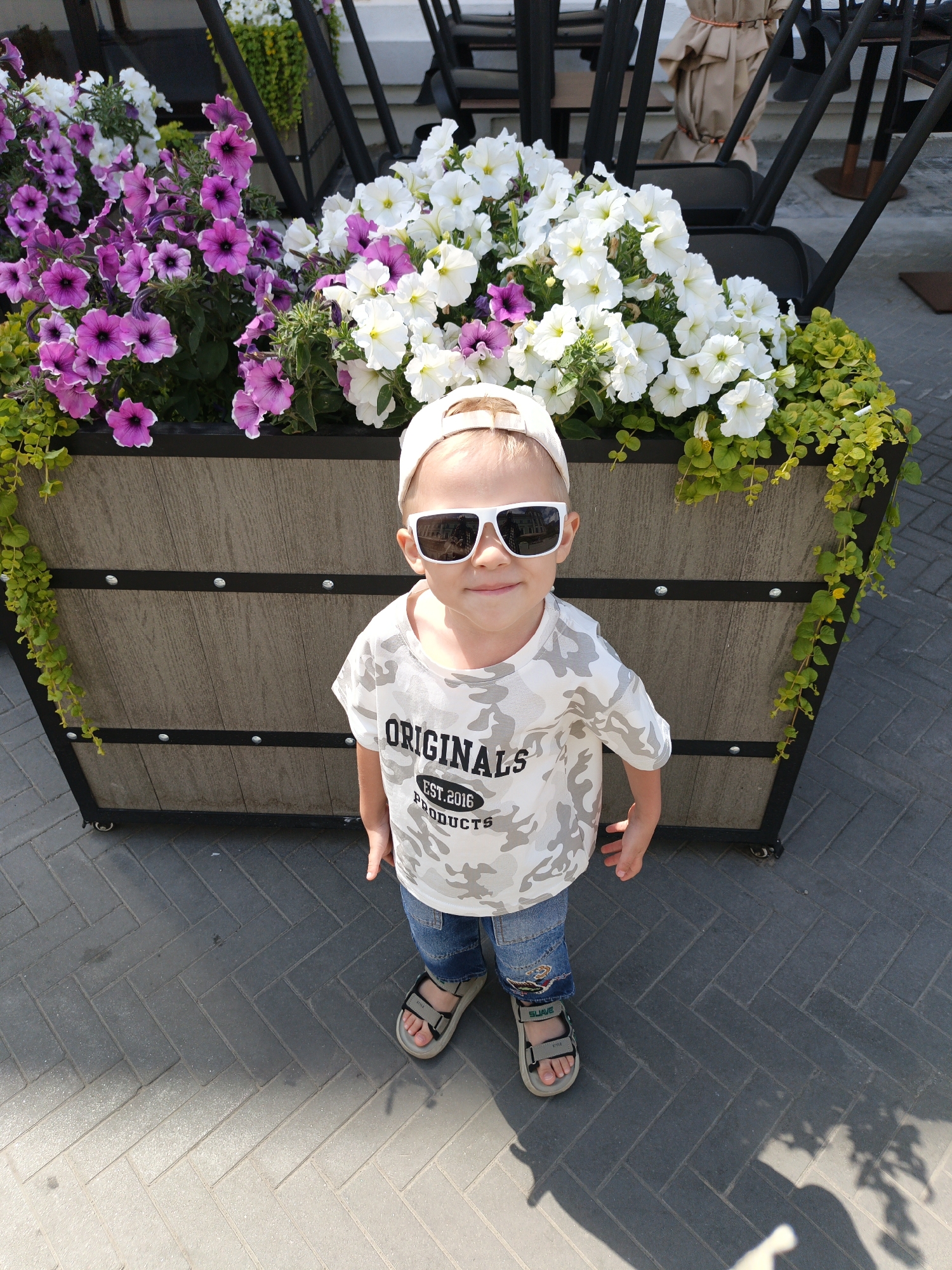 A young child wearing a camouflage t-shirt, blue jeans, sunglasses, and sandals stands in front of a large planter box filled with pink and white petunias. The planter box is made of a wood-like material with black metal accents and sits on a paved surface. In the background, stacked chairs and a covered object suggest an outdoor seating area, possibly at a cafe or restaurant in Kazan, Russia. The lighting indicates a sunny day, with shadows cast on the ground. The child is looking towards the camera with a slight smile.