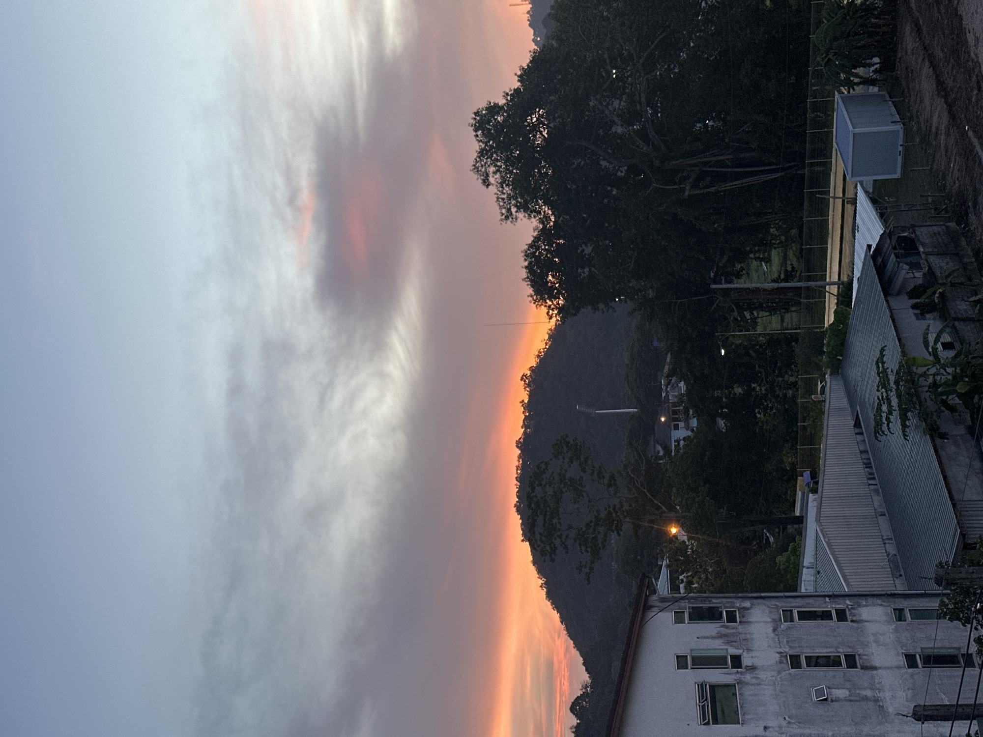 The image depicts a natural landscape at dusk, with a prominent hill in the background silhouetted against a vibrant sky painted with hues of orange, pink, and blue. The sky displays wispy clouds, suggesting the final moments of sunset. In the foreground, a multi-story building with numerous windows stands on the left, its facade appearing somewhat weathered. Adjacent to it are several low-rise structures with corrugated metal roofs. Dense foliage, including large trees, dominates the middle and right portions of the image, partially obscuring the background hill. A tall, slender tower, possibly a light pole or communication mast, is visible on the hill. The overall setting appears to be a semi-urban or suburban area, with elements of both built structures and natural vegetation. The time of day suggests an evening scene.