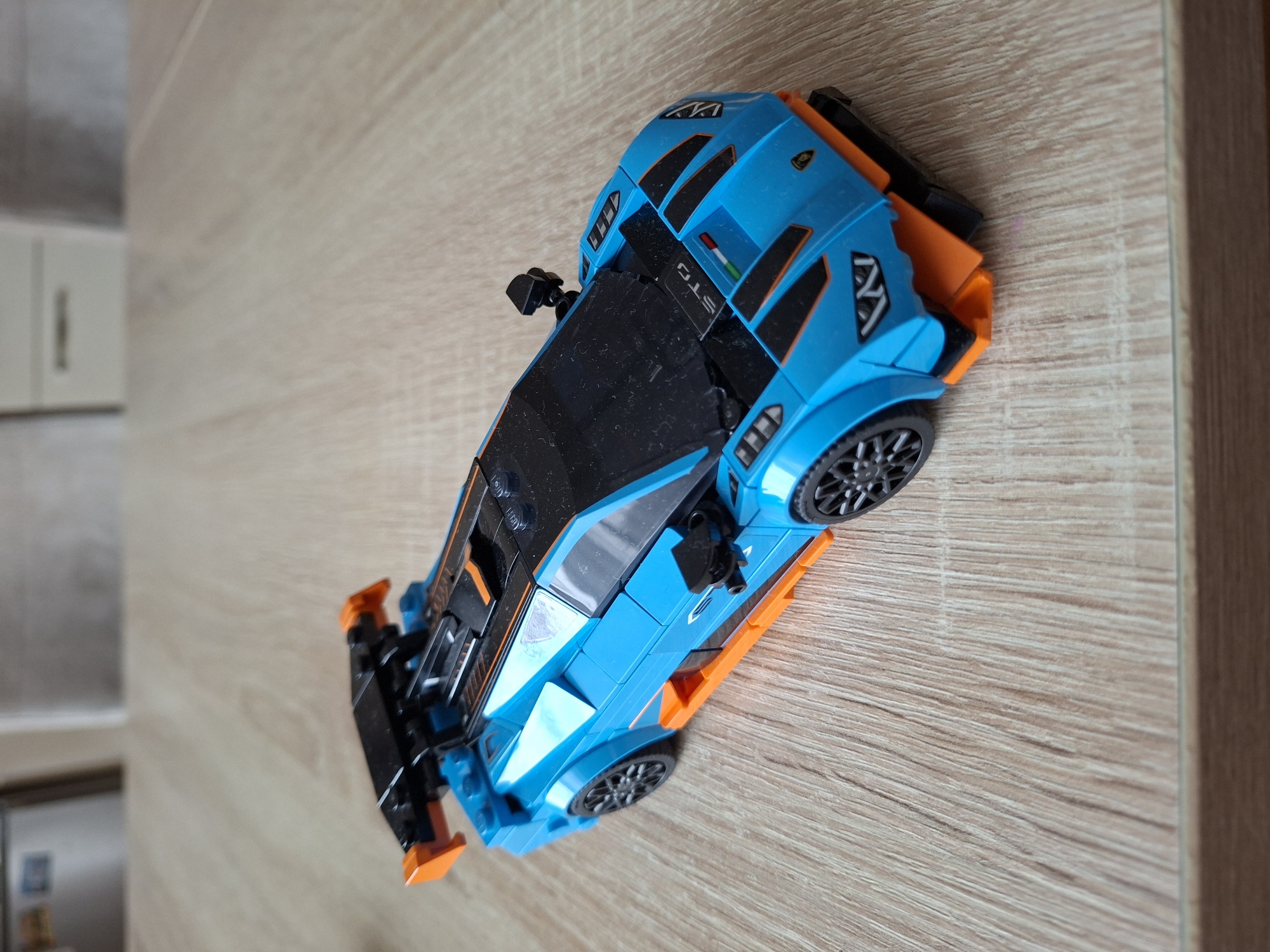 A blue and orange toy Lamborghini sports car is positioned on a light brown wooden surface. The car is angled, facing towards the right side of the frame and slightly away from the viewer. The car features black accents on its body, including a large rear spoiler and detailing on the hood and sides. The wheels are black with intricate spoke designs. The surface it rests on appears to be a table or desk with visible wood grain. The background is blurred, showing hints of light-colored furniture or appliances. There is no indication of the city of Baden-Baden, Germany, in this image.