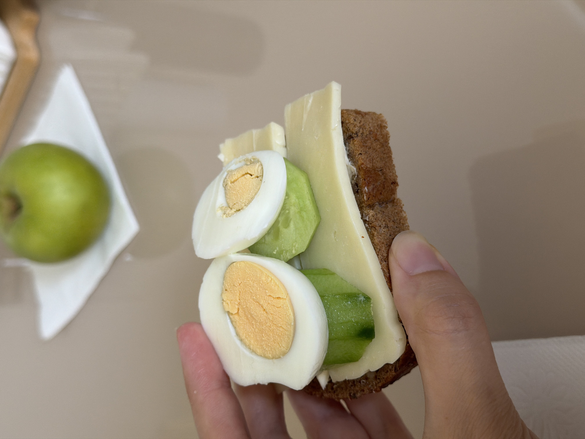 A person's hand is holding a slice of dark bread topped with cheese, cucumber, and sliced hard-boiled eggs. In the background, a green apple sits on a white napkin. The scene appears to be indoors, likely on a table. There is no information available to determine the location in Izhevsk, Russia.