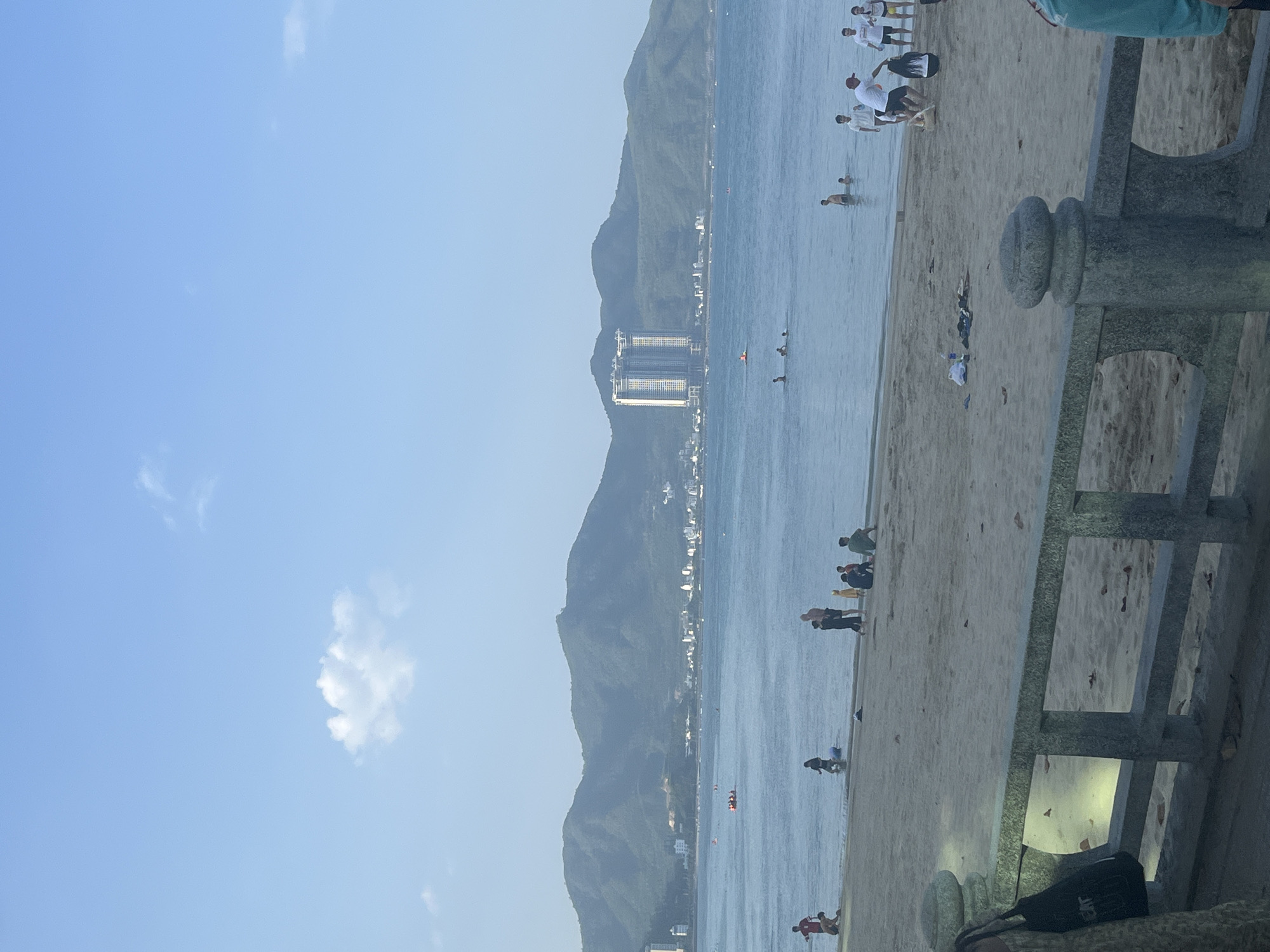 The image shows a beach scene in Nha Trang, Vietnam, with the Nha Trang Bay in the background. A long stretch of sandy beach is visible in the foreground, bordered by a stone railing. Several people are present on the beach, some sitting and others wading in the shallow water.
In the middle ground, the blue waters of Nha Trang Bay are calm, with a few small boats scattered across the surface. Further in the background, lush green mountains rise up, with a strip of buildings along the coast, indicating the urban development of the city. Two tall, modern buildings, likely hotels or condominiums, stand out prominently against the mountainous backdrop. The sky is a clear, pale blue with a few wispy clouds. The overall impression is a serene coastal landscape with recreational activities taking place.