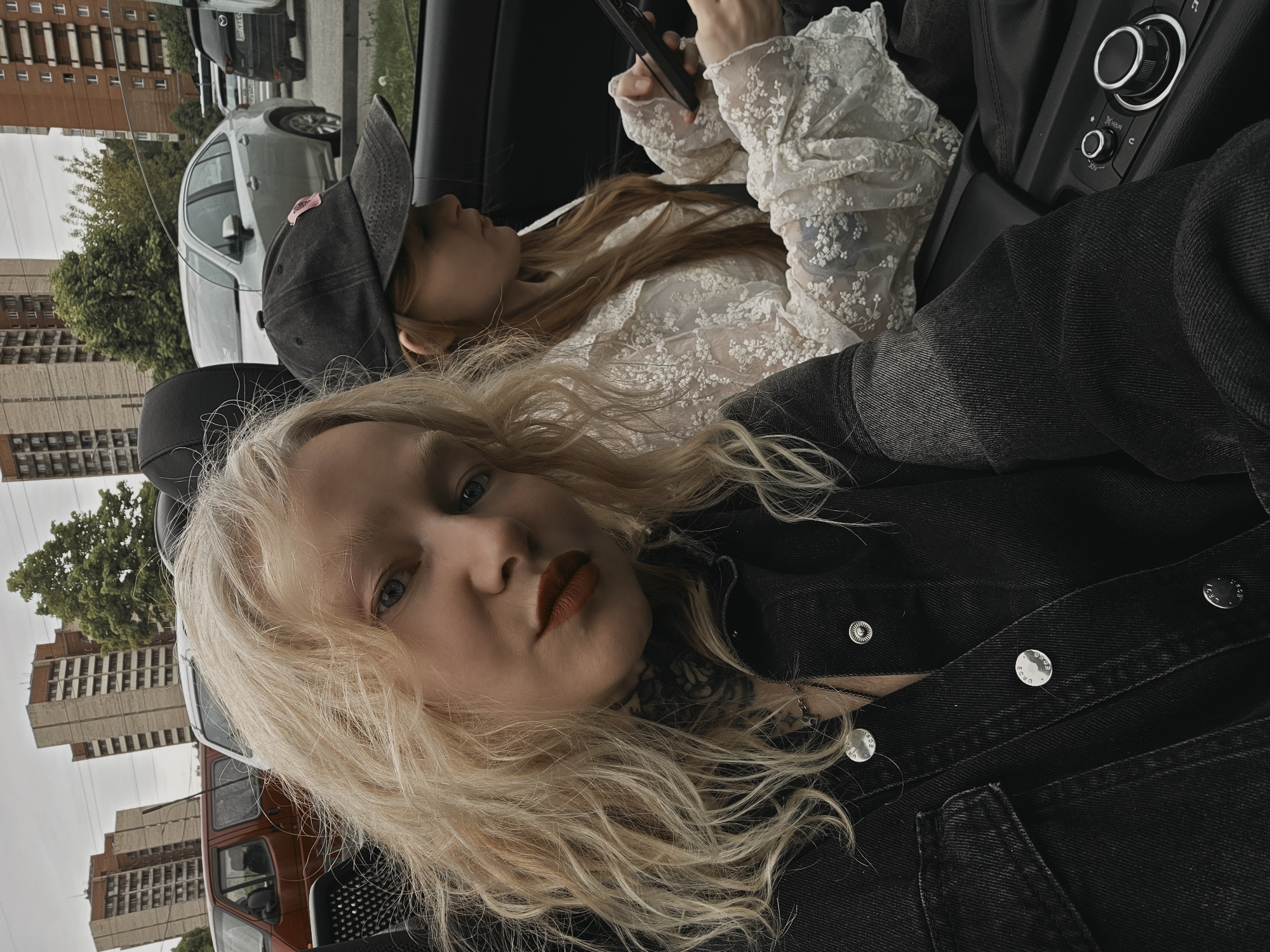 Two young women are in a car, angled such that the viewer is looking upward slightly at them. The woman in the foreground has light blonde, wavy hair, blue eyes, and is wearing a dark denim jacket. A visible tattoo is on her neck. The woman in the background has auburn hair, is wearing a baseball cap, and a white lace top, and is looking at a phone in her hands. Part of the car's console is visible, along with exterior buildings, including what may be a bus or larger vehicle and apartment buildings. Considering the architectural styles and vehicles, the location is most likely Saint Petersburg, Russia.