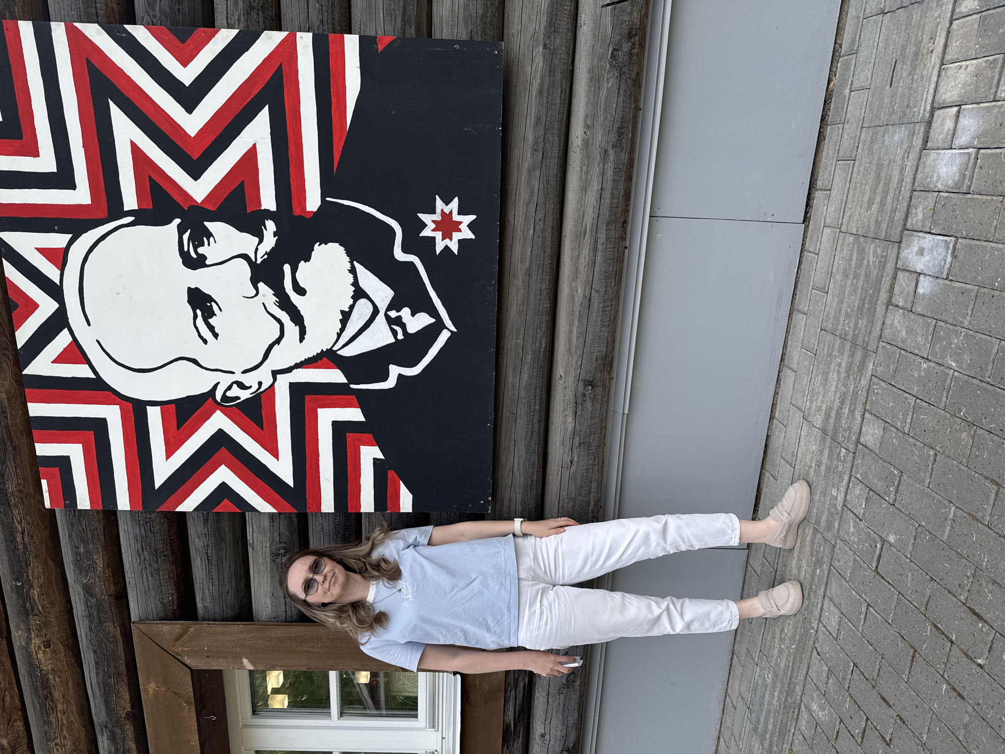 A woman stands in front of a wooden building in Votkinsk, Russia. She wears a light blue t-shirt, white pants, and light tan shoes. She also wears glasses. A poster of Vladimir Lenin is mounted on the side of the building. The poster is painted in black, white, and red. A window is visible on the lower left of the frame. The ground is paved with gray bricks.