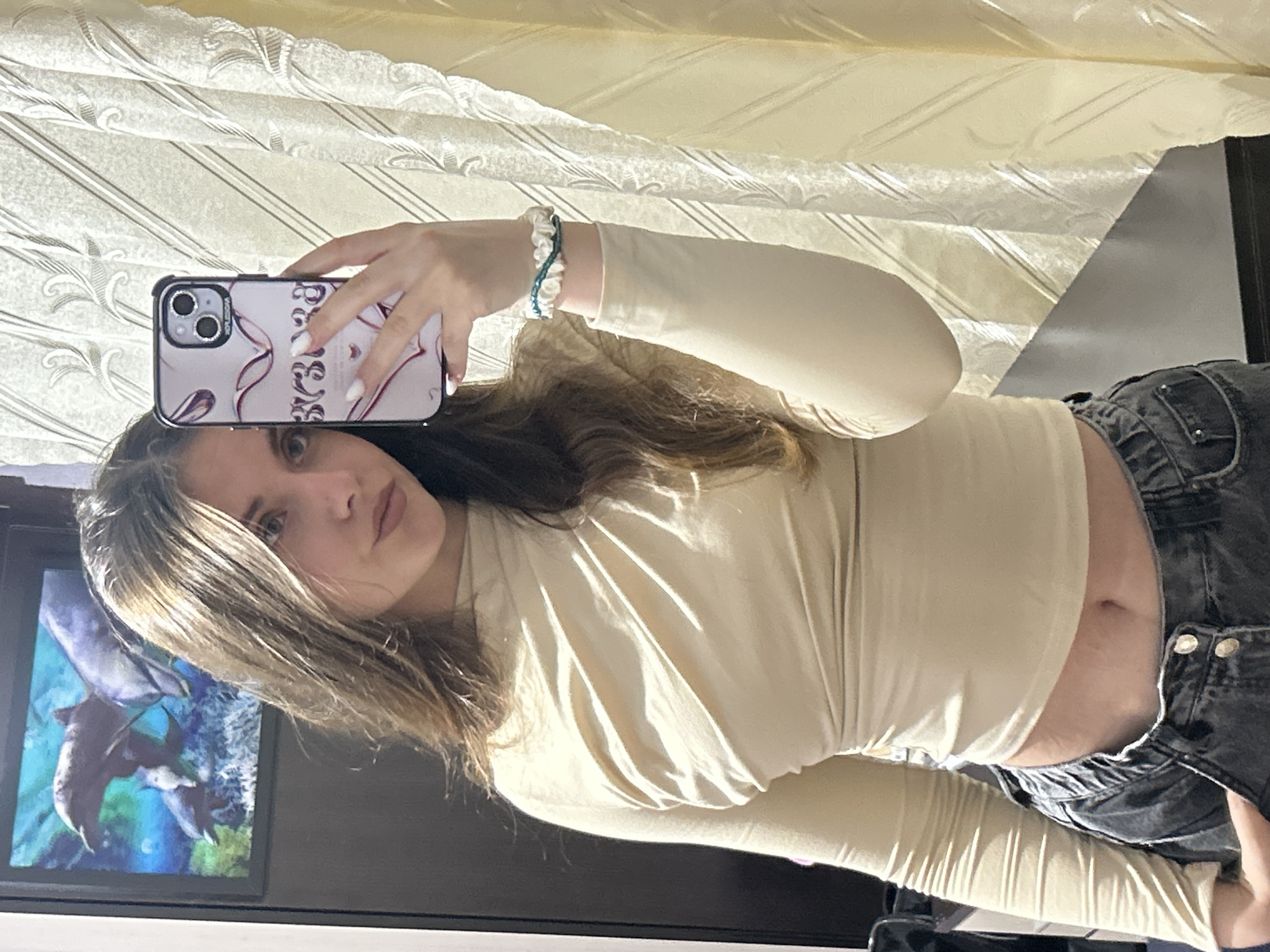 A young woman with long, brown hair takes a selfie in a room. She is wearing a light beige, long-sleeved, draped top and dark-wash, low-rise jeans. She has on a white bracelet with teal accents and a ring on her left hand. Her phone, which has a pink and white case with a heart design, is held in her right hand. In the background, a framed picture of dolphins is visible on a dark wall, and a yellow, patterned curtain hangs to the right.