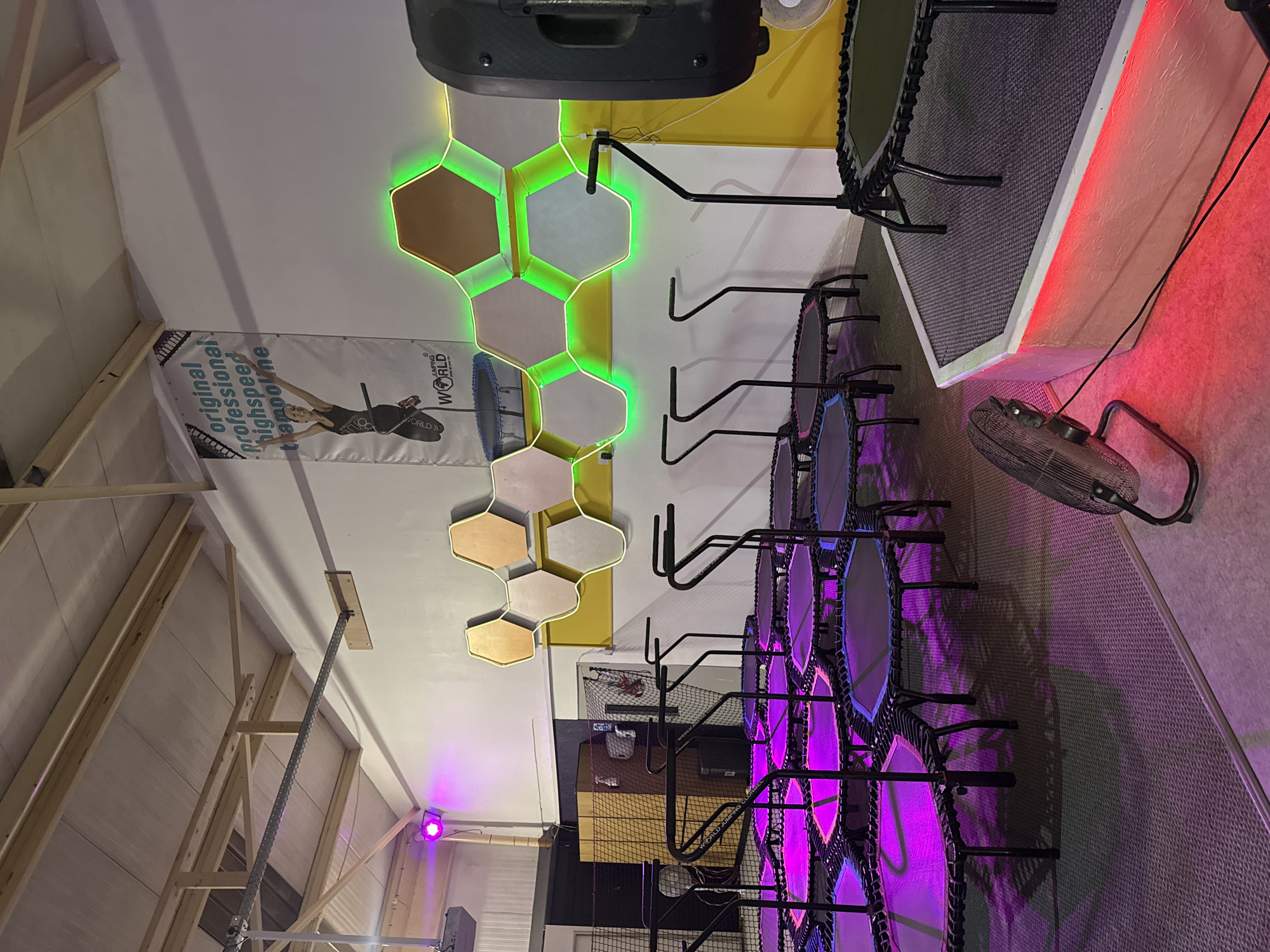 The image depicts an indoor trampoline park. The scene is dominated by numerous hexagonal trampolines arranged in a grid, illuminated by purple and pink lighting. Each trampoline is equipped with black safety handles. In the background, a banner hangs from the wall, featuring a woman jumping and the text "original professional highspeed trampoline". The wall is decorated with honeycomb-shaped panels outlined in green neon lights. A large speaker is visible on the right side of the image, positioned on a raised platform with red lighting at its base. A black oscillating fan is situated on the floor in the foreground. The environment suggests a recreational facility designed for fitness or entertainment. There are no individuals visible in the image. The location is identified as Clonmel, Ireland, suggesting this is a business or venue within that town.