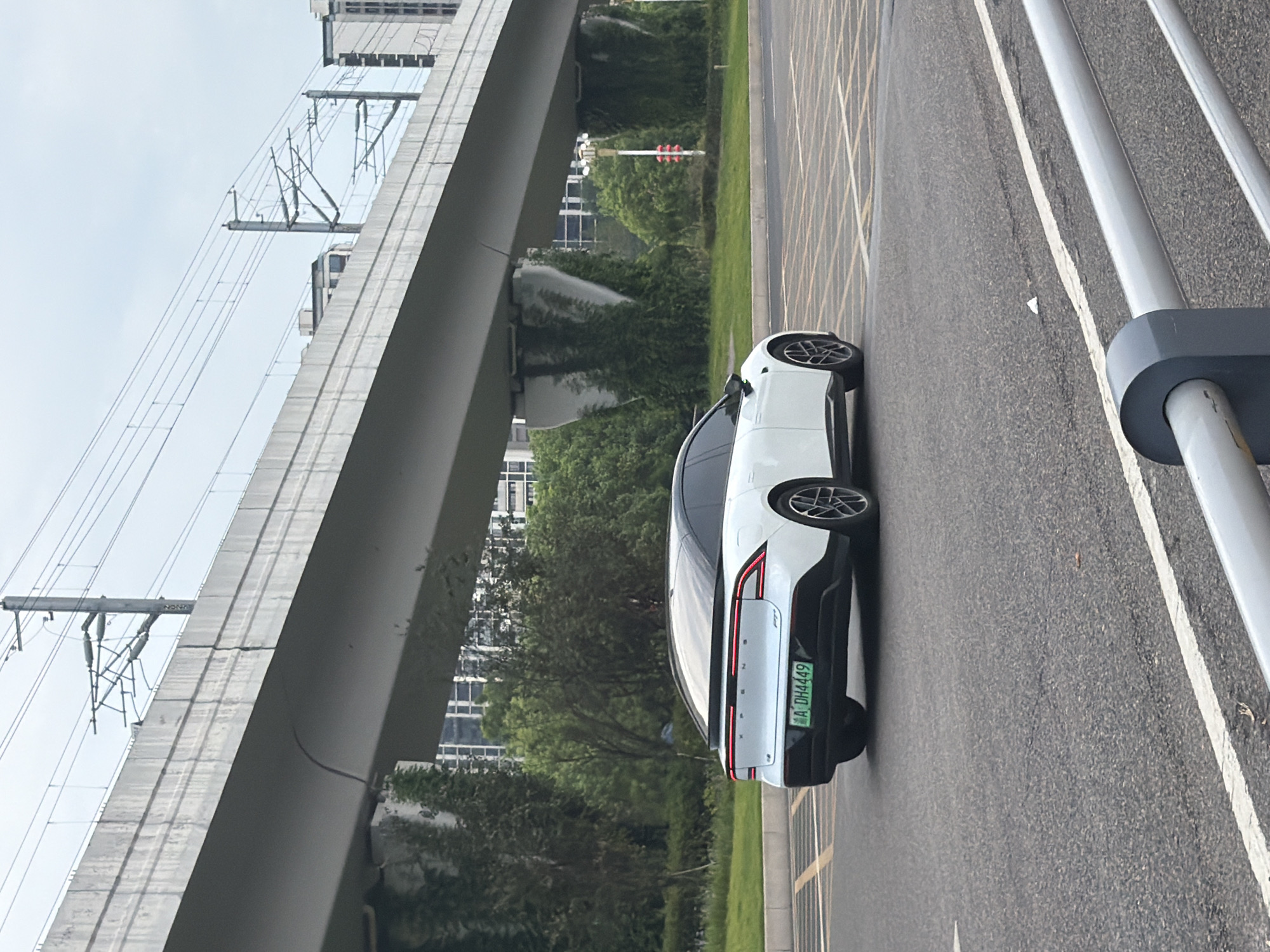 A white electric car with a black roof is driving on a road in Jiangjin, China. The car has a license plate that reads "A DH4449". The car is under an elevated highway or overpass, with buildings and trees in the background. The sky is overcast. The road has yellow and white markings.