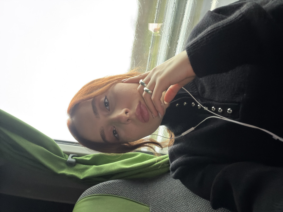 The image captures a young individual, female, with red-brown hair, seated within a vehicle, reportedly in Moineville, France. She is wearing a black long-sleeved top featuring a mock-neck and a front placket adorned with silver-colored eyelets or snaps. White wired earphones are visible around her neck and leading to her ears. Her left hand is positioned near her chin, displaying two silver-colored rings on her ring finger. To her left, part of a grey fabric seat and a green curtain are visible. To her right, a window shows a blurred exterior with an overcast sky and streaks of rain or moisture on the glass.