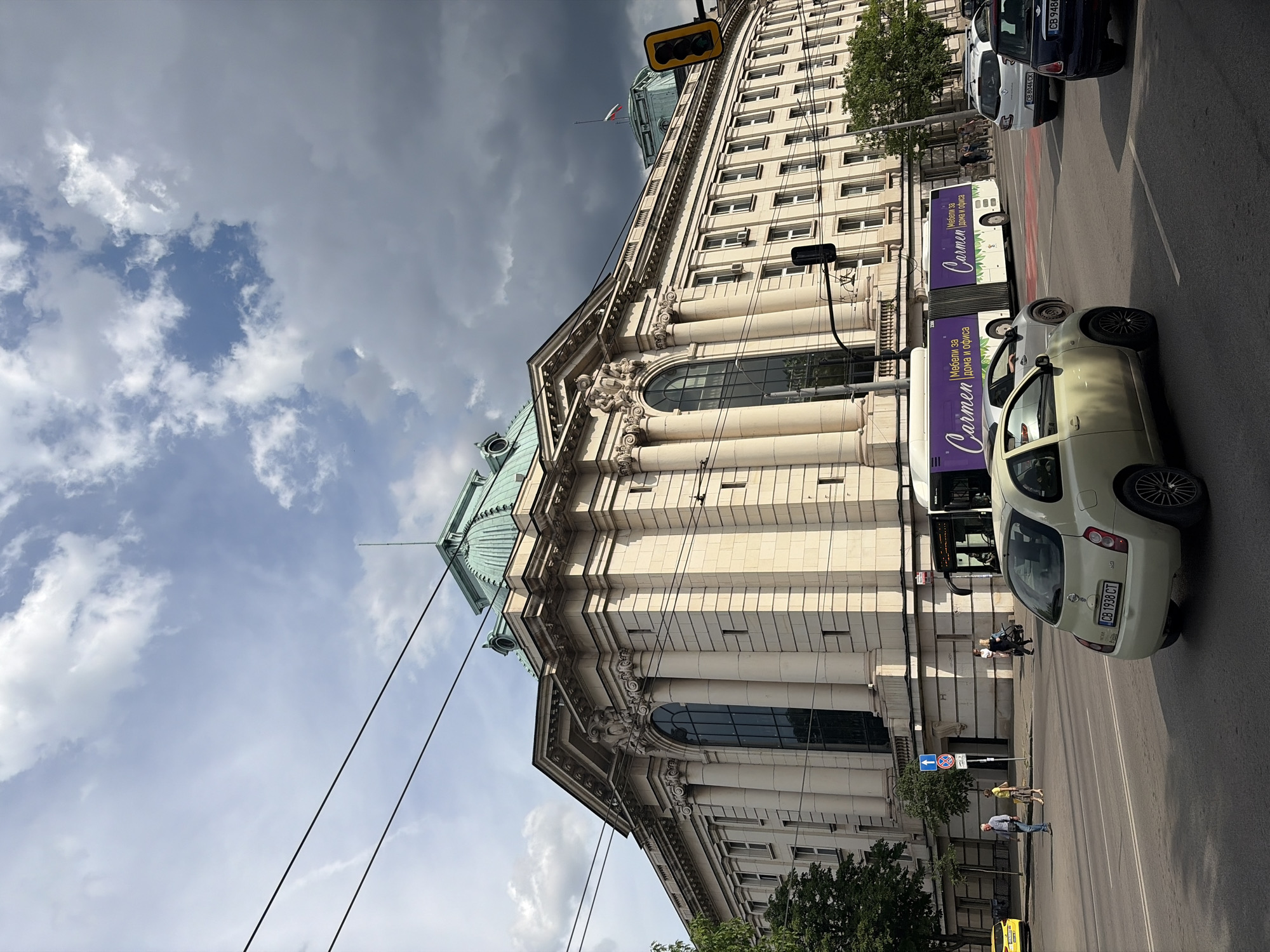 This image captures a view of the iconic National Assembly building in Sofia, Bulgaria. The imposing sandstone structure features a prominent green domed roof and classical architectural elements like columns and ornate carvings. In the foreground, a light-colored Nissan Micra is stopped at a traffic light. Behind it, a bus with the "Carmen" logo, advertising furniture for home and office, is also visible. Several other cars are present on the road, and pedestrians are walking on the sidewalk. The sky is partly cloudy, with dramatic dark clouds gathering in the upper right portion of the image, juxtaposed against brighter sections of blue sky. Overhead, power lines crisscross the scene, common in urban environments. The overall impression is a street-level view of a significant landmark in a bustling city.