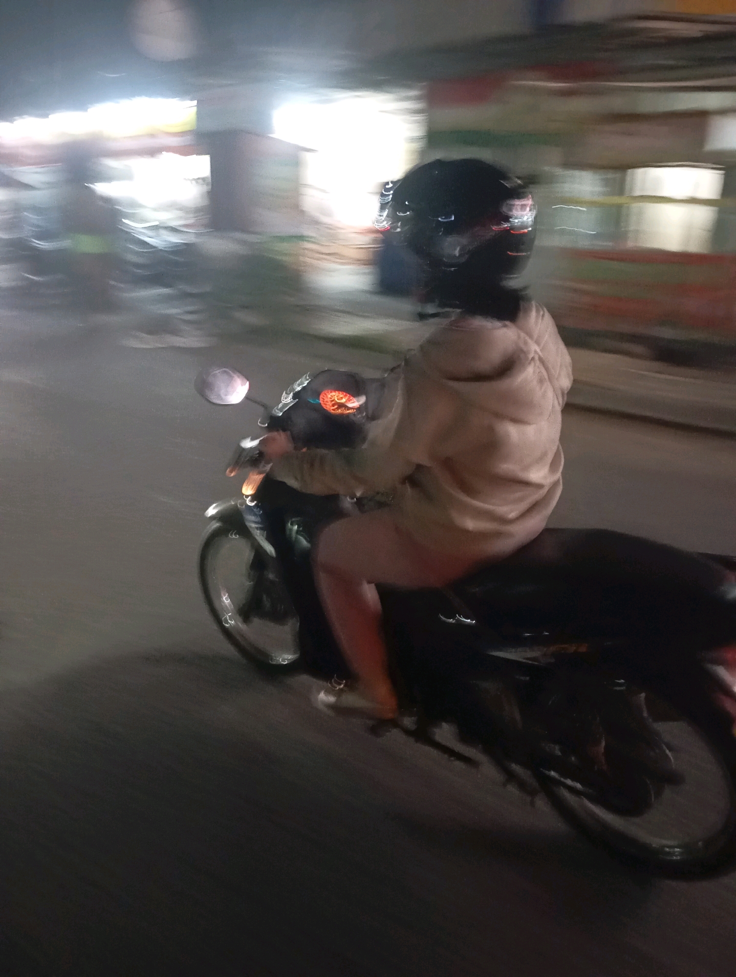 A person wearing a black helmet and a light-colored hooded jacket rides a motorcycle at night on a paved road in Babelan, Indonesia. The image is motion-blurred, indicating movement. The background shows dimly lit structures and possibly other vehicles. The rider's legs are visible, and they are wearing light-colored pants. The motorcycle is dark-colored, and its wheels are also blurred due to motion. The scene appears to be on a street with overhead lighting and some ambient light from buildings.