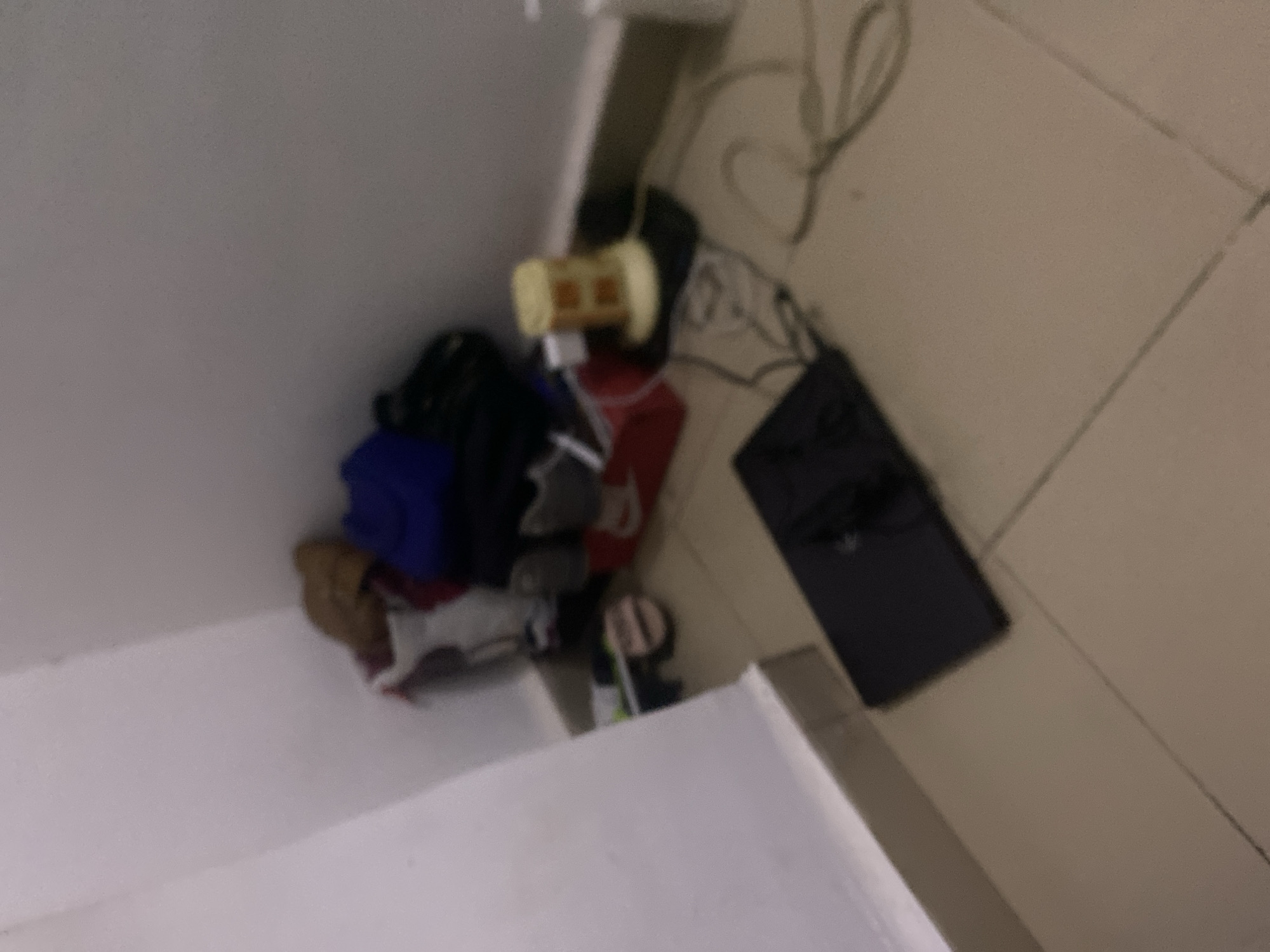 The image depicts a cluttered corner of a room, possibly a storage area or a less-used space. The floor is tiled with light-colored tiles, and the walls are painted white. In the corner, a pile of items includes what appears to be a brown bag or piece of fabric, a dark blue garment, and possibly some shoes or footwear. Adjacent to this pile is a red box, with white and red markings suggesting it might be a shoe box. On top of the red box, there are some gray and white items, possibly shoes or bags.
Further to the right, a yellow object with square-shaped cutouts sits on a black surface, which is connected to various cables and wires scattered on the floor. In the foreground, a black laptop or similar electronic device is placed on the floor, with some dark objects on top of it. The overall impression is one of disorganization and accumulated belongings. The scene does not provide any specific visual cues to pinpoint its location within Howayi, Nigeria.