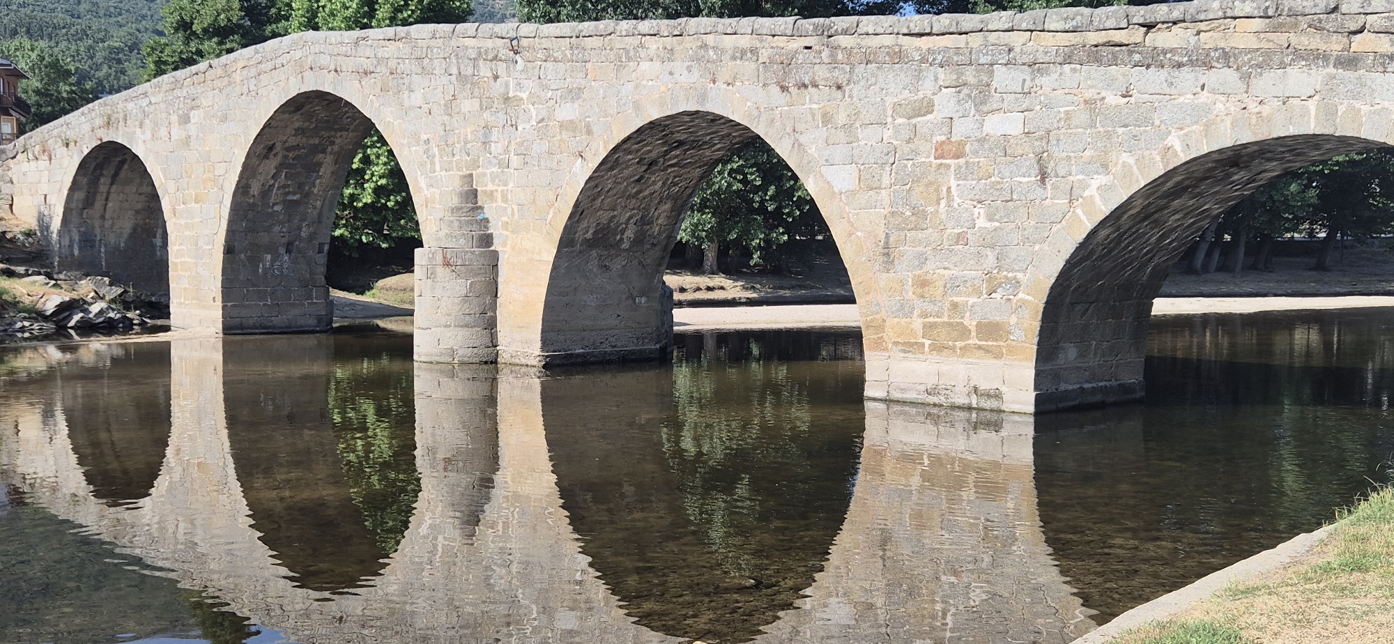 A wide, stone arched bridge spans a calm body of water, likely a river, with its reflection clearly visible on the surface. The bridge is constructed from light-colored, roughly hewn stones, forming five prominent arches, though only three are fully visible from this angle. The left side of the bridge is partially obscured by trees and a building with a reddish-brown facade. The right side of the bridge extends out of frame. The water is dark and still, reflecting the bridge and the lush green trees that line the far bank. The lighting suggests a sunny day, with shadows cast by the arches onto the water. The immediate foreground on the right features a grassy embankment. No people are present in the scene.