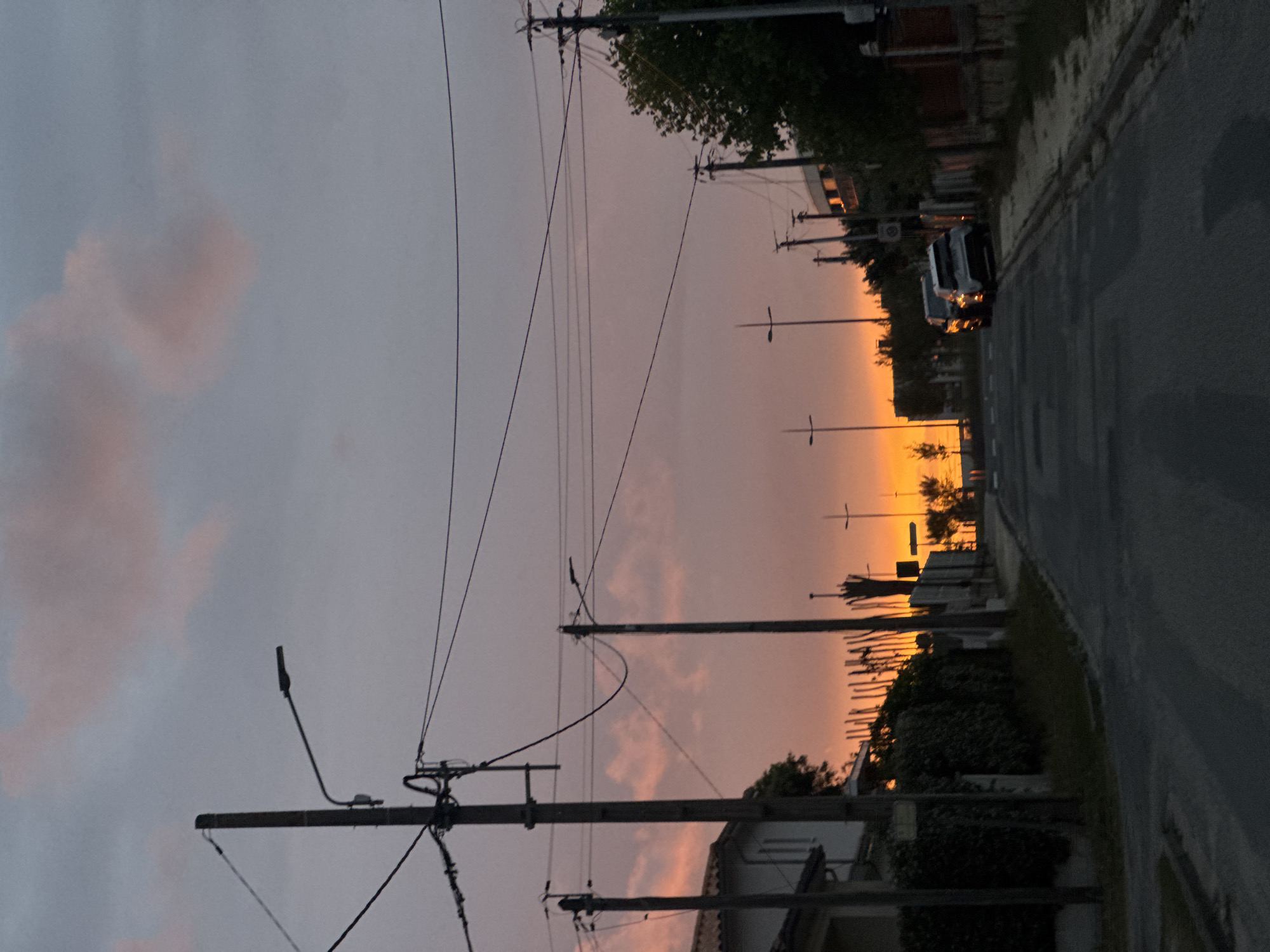 The image shows a street in Soulac-sur-Mer, France, at dusk. A row of telephone poles lines the left side of the street, with wires crisscrossing the sky. On the right, a narrow asphalt road stretches into the distance, bordered by a curb and vegetation. Buildings and parked cars are visible on the right, silhouetted against the orange glow of the setting sun. The sky is a mix of grey and pink hues, with darker clouds on the left and brighter colors towards the horizon.