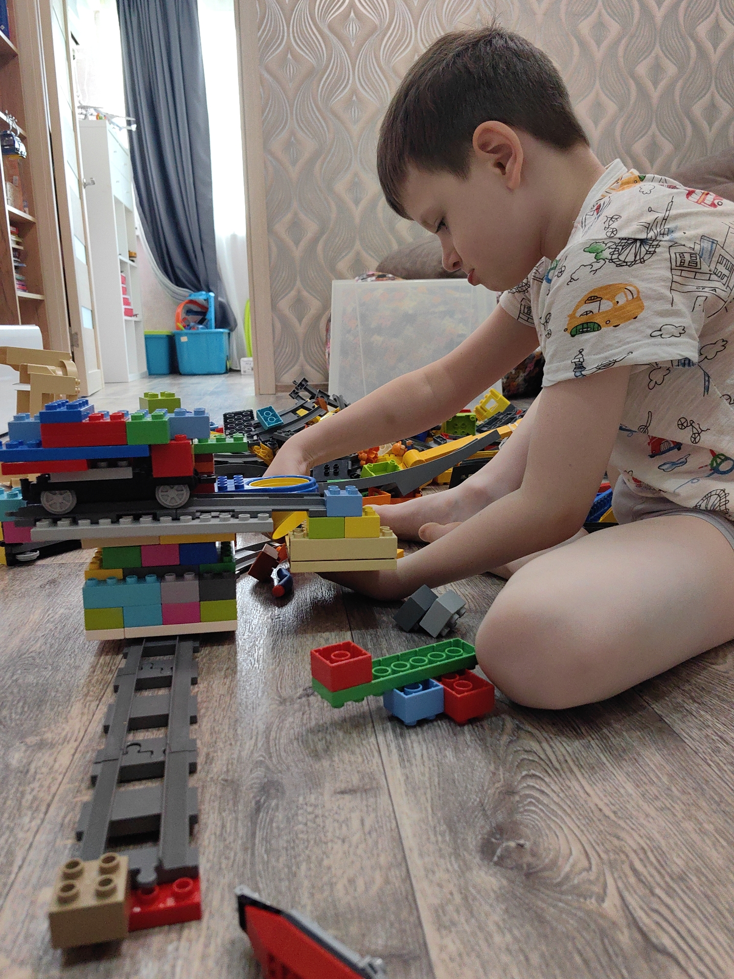 A young boy is sitting on a wooden floor, playing with colorful building blocks. He is wearing a white t-shirt with a pattern of cartoon vehicles. The boy is focused on constructing a train or a similar structure made of various colored blocks, including red, blue, green, yellow, and gray. Some of the blocks are stacked on top of each other to form a train car and a track. In the background, there are more building blocks, a bookshelf, a blue storage bin, and a gray curtain. The room appears to be a child's playroom or bedroom. The image does not provide any information about the city of Sochi, Russia, or any other specific location.