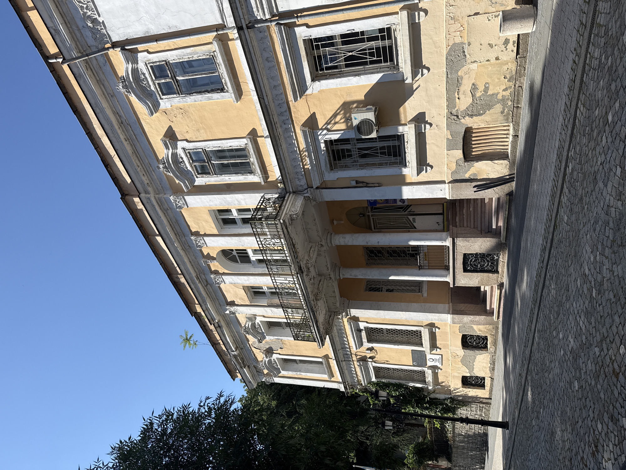 This image depicts the exterior of a historic, aged building with pale yellow stucco walls, contrasting with white architectural details. The building appears to be several stories high, featuring numerous windows, some with decorative metal grilles. An air conditioning unit is mounted on the facade. A balcony with an ornate black metal railing extends across a section of the building. The building's architecture suggests a European style, possibly from the 19th or early 20th century.
To the right of the building, a cobblestone street runs alongside it, with shadows cast on the pavement, indicating it is daytime with the sun positioned at an angle. Greenery, including trees and ivy, grows around the base of the building and along a stone wall.
The scene is set in Plovdiv, Bulgaria. The clear blue sky suggests fair weather. There is no discernible activity or event taking place, and no people are visible. The overall impression is one of a timeless, somewhat weathered, urban environment. There is some faint, unreadable text on a plaque on the building and some signs visible through the windows.