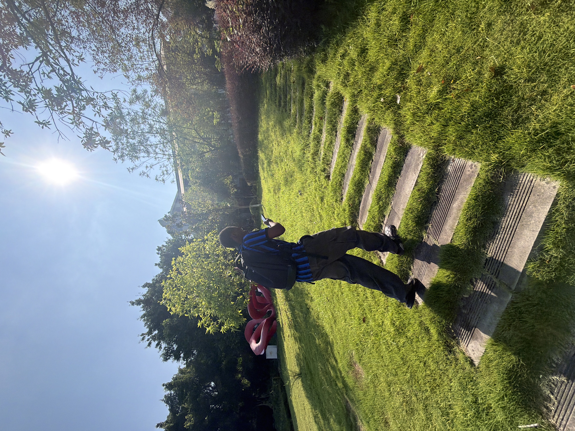 A person with a backpack is walking up a grassy slope with wooden steps in Jiangjin, China. The sun is shining brightly, casting shadows. There is a red sculpture in the background. The environment appears to be a park or a garden. The person is wearing a blue and black striped shirt and dark pants.