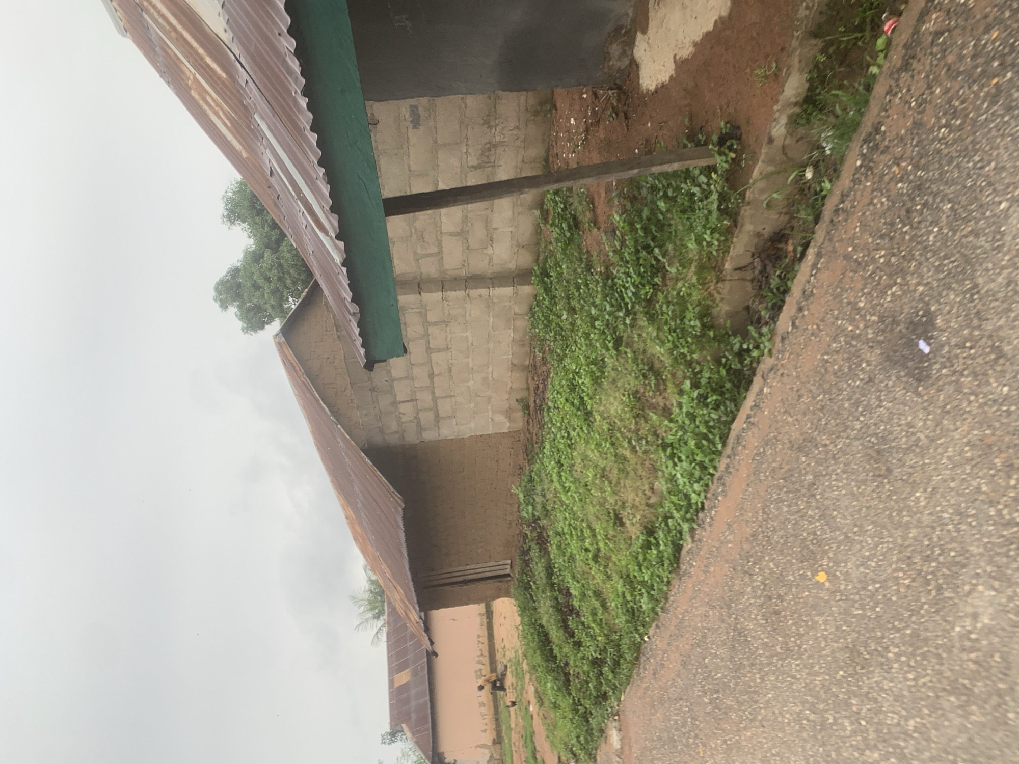 In the city of Serti, Nigeria, a dirt road borders a residential area featuring multiple buildings with corrugated metal roofs. In the foreground, the road is asphalted and shows some scattered debris. To the right of the road, a narrow concrete drainage ditch is visible, overgrown with weeds and containing various pieces of trash.
Beyond the ditch, a building constructed with light-colored concrete blocks is prominent, with a section of its rusty corrugated metal roof extending over a green overhang supported by a wooden post. To the left of this structure, another building made of mud or earth walls is partially visible, also topped with a corrugated metal roof. Further to the left, a third building with a light-colored facade and a corrugated metal roof can be seen.
In the yard between the buildings, a person in a yellow shirt is seated, accompanied by a chicken. The ground in front of the houses is covered with green vegetation and patches of red soil. The sky is overcast and cloudy, suggesting a potentially dull or rainy day.