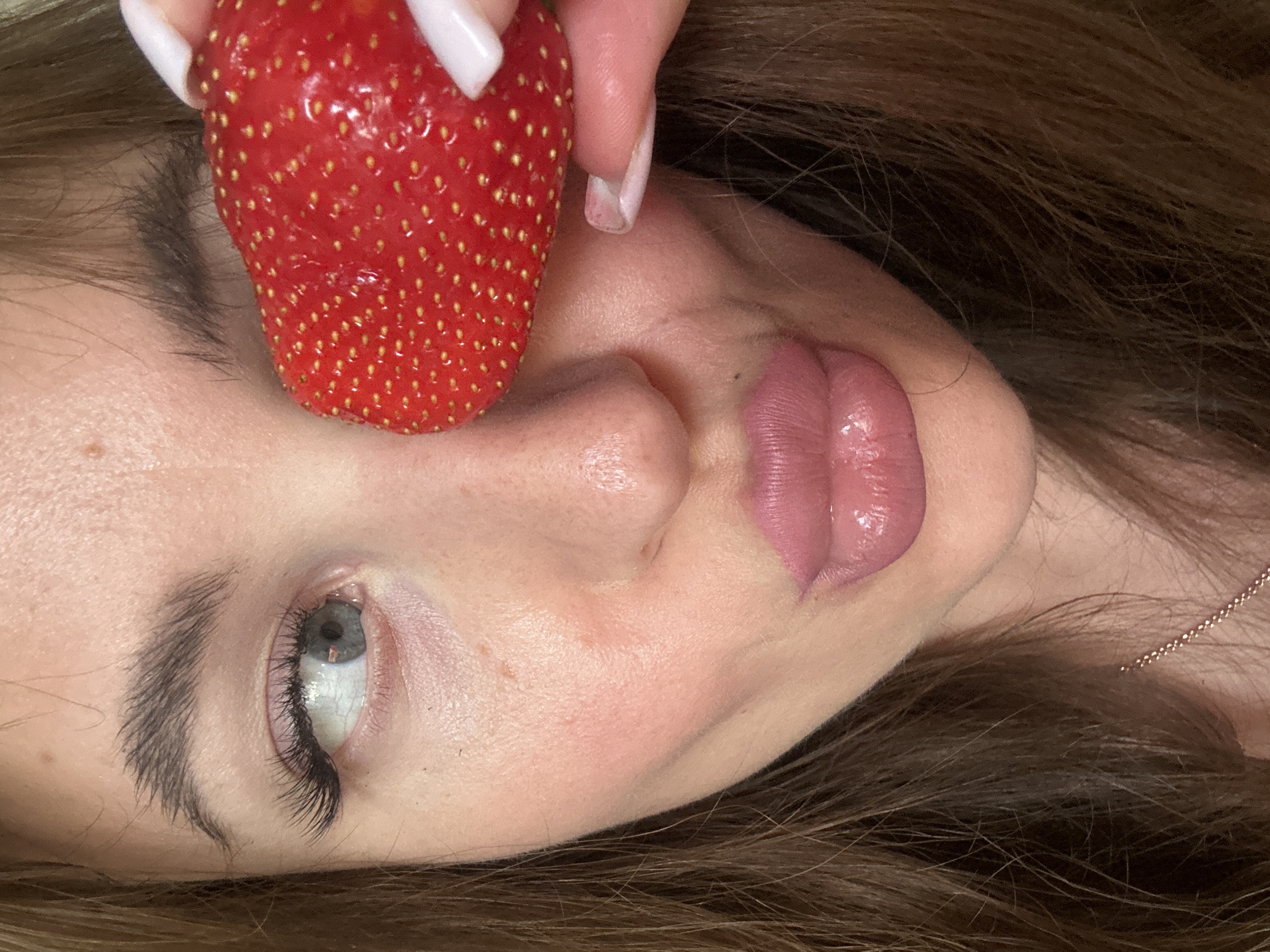 A strawberry is held near the upper portion of a woman's face. The woman is lying on her side, and the strawberry is held in front of her eye and nose. The woman has long brown hair, fair skin, and pink lips. She is wearing a thin, gold necklace. The background is out of focus. There is no indication of this photo being taken in Chaikovsky, Russia.
