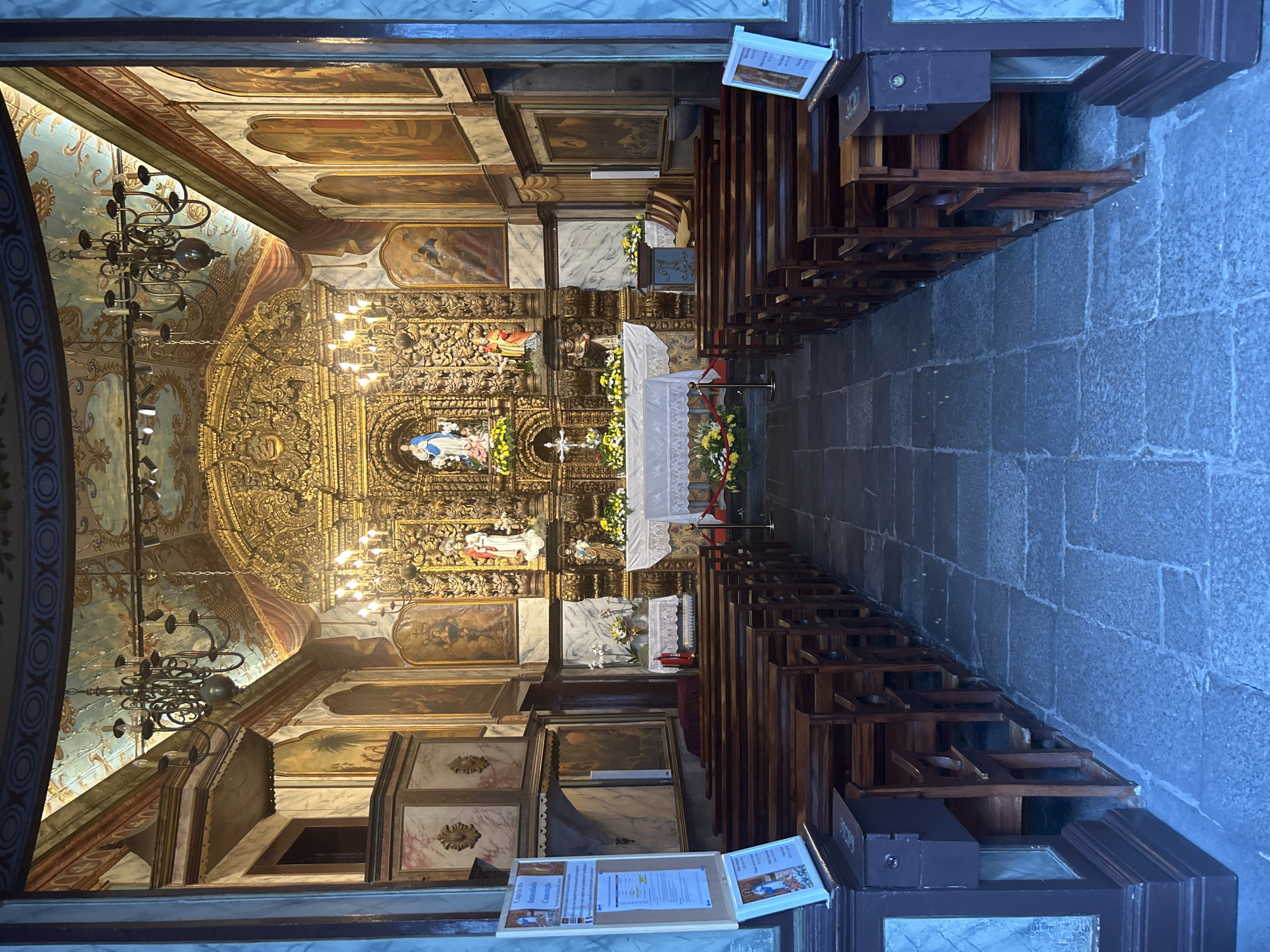 The image depicts the interior of the Capela da Nossa Senhora da Conceição in Câmara de Lobos, Portugal. The chapel features ornate gold detailing on the altar and upper walls, with paintings adorning the walls. Wooden pews line either side of the stone-paved aisle, leading towards the altar. Chandeliers hang from the painted ceiling. Information posters are present near the entrance. No people are visible.