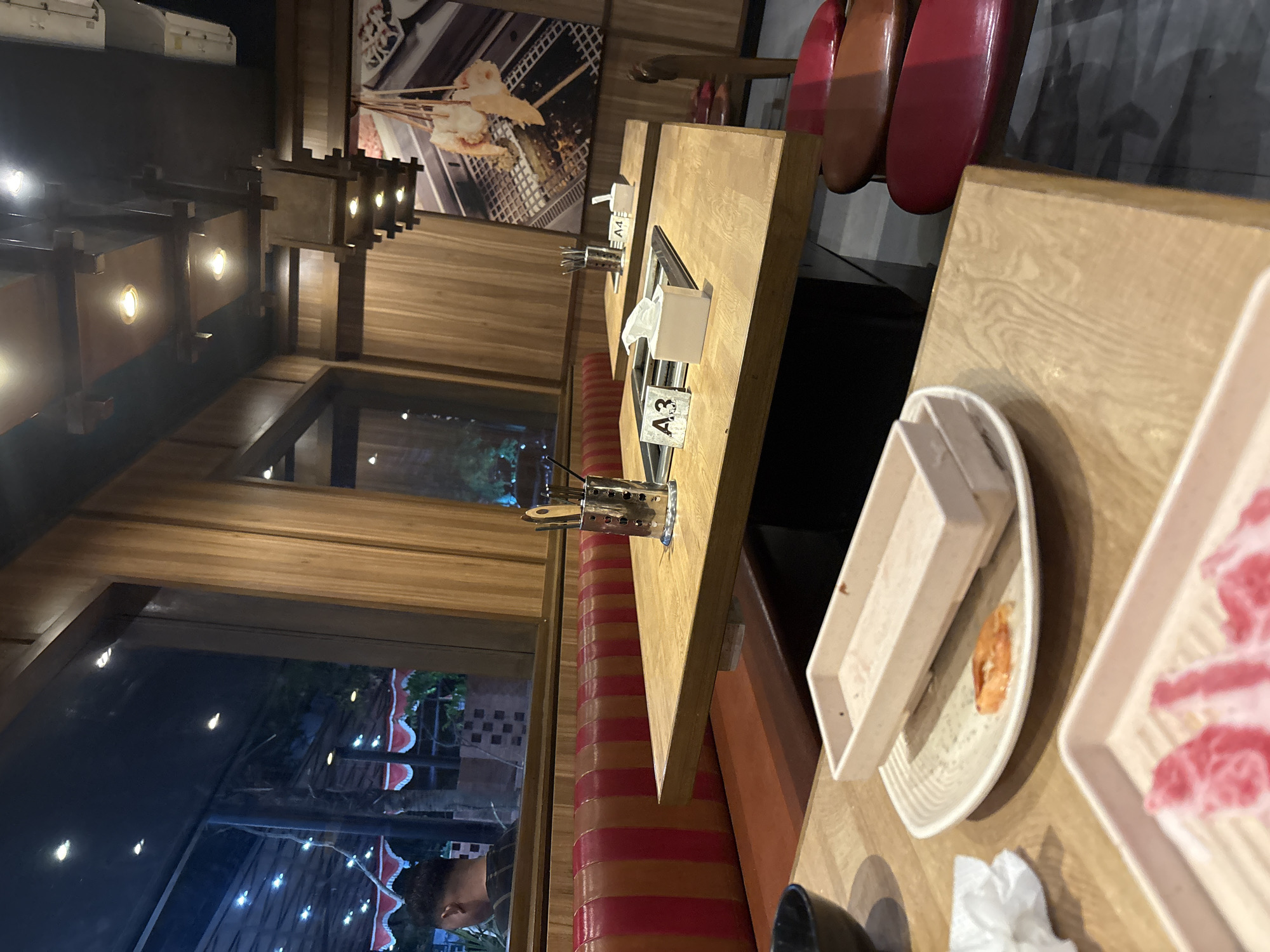 The image depicts an interior dining scene at a restaurant, likely a Korean BBQ or similar establishment, in Cirebon, Indonesia. The setting is casual and modern, with wooden tables and paneling, plush booth seating in red and orange stripes, and overhead lighting fixtures integrated into wooden structural elements.
Two wooden tables are prominently featured. The table in the foreground has a white tray with a single cooked shrimp on a round plate. Next to it is a rectangular white tray and a crumpled white napkin. The table behind it displays a metal utensil holder with tongs and chopsticks, a white box of tissues, and a table number sign labeled "A4". A similar table number sign, "A3", is visible on the table in the foreground, also with a metal grilling apparatus and a tissue box.
In the background, through a large window, a nighttime scene is visible with string lights and red and white decorative awnings, suggesting an outdoor or semi-outdoor seating area. A person is partially visible in the reflection of the window, looking outwards.
The overall ambiance suggests a dining establishment prepared for patrons, with food items and place settings arranged on the tables. The image is taken from a slightly elevated angle, looking down at the tables. The lighting is warm and ambient.