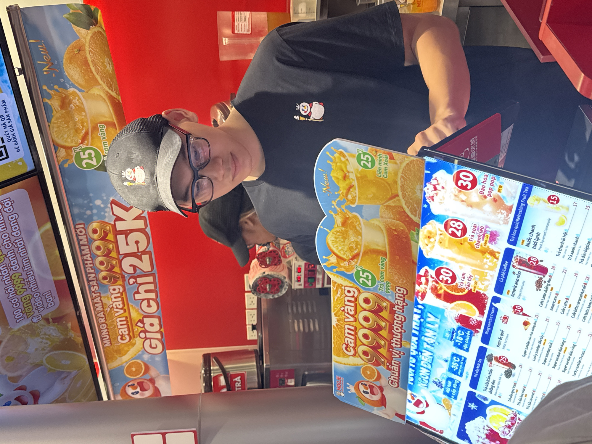 Here's a matter-of-fact description of the image:
**Scene:**
The image captures an indoor scene, likely within a Mixue beverage shop, evident from the branded signage.
**People:**
A young man with glasses and a baseball cap is centrally positioned. He appears to be looking at the camera. Part of another person's head is visible behind him.
**Subjects:**
The focus is on the Mixue menu and promotional material, showcasing images and prices of various drinks, including "Cam Vàng" (orange juice) and other fruit teas. The signage also includes Vietnamese text and branding.
**Objects:**
Several Mixue menu boards, a point-of-sale tablet or screen, drink preparation equipment, cups, a tea dispenser, and a counter area are present.
**Actions:**
The man seems to be working or positioned near the service area of the beverage shop. He is looking at the camera.
**Interactions:**
The interaction is between the person and the camera lens. The menu boards and pricing suggest customers would be interacting with the menu to order drinks.
**Environment:**
The shop's decor includes a red wall, contributing to a brightly lit and branded environment. The overall setting conveys a typical commercial space within a city. The Vietnamese text indicates this is in Vietnam, possibly Hà Nội (based on the OCR results on the image).