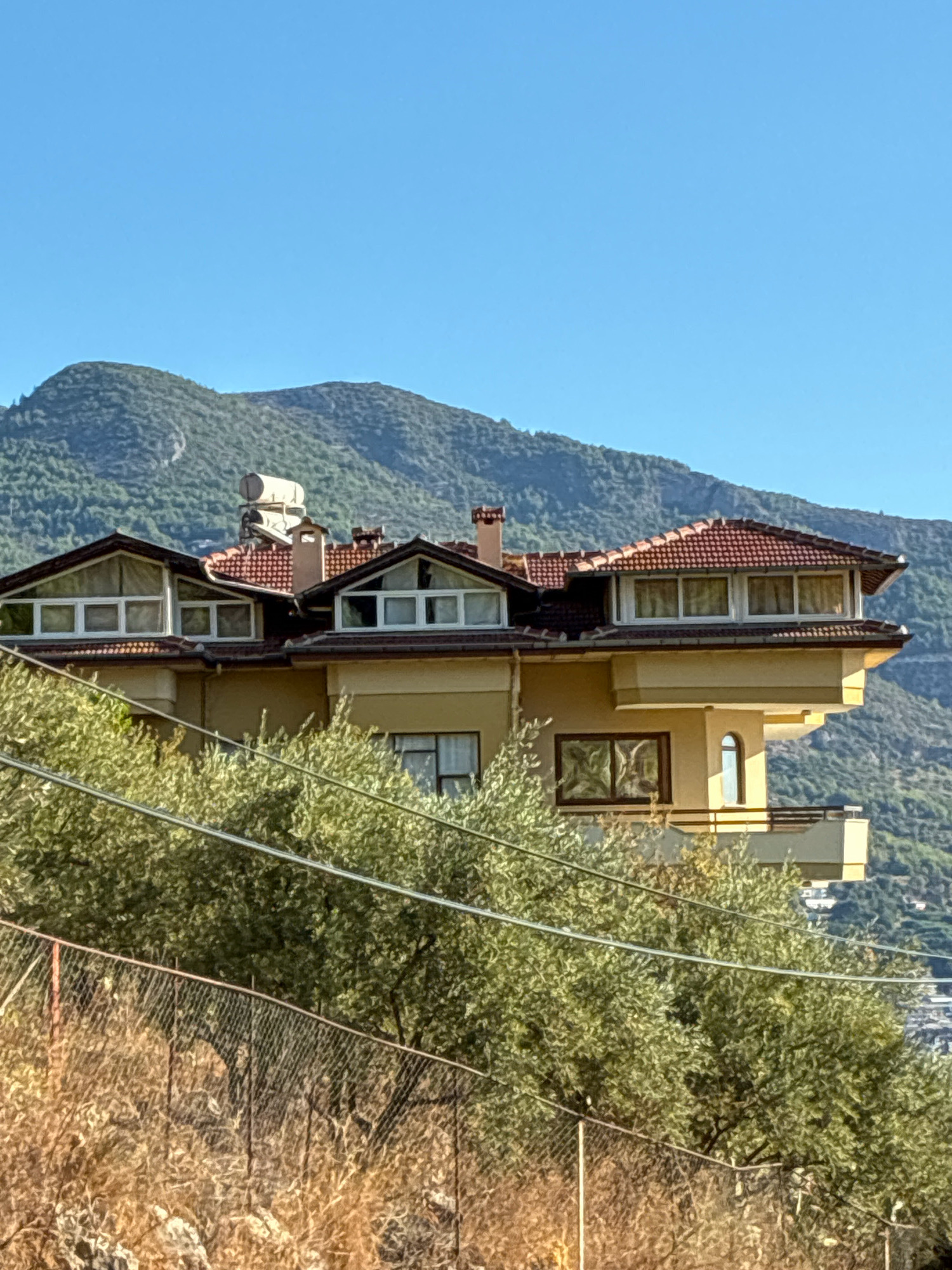 This is a photograph of a multi-story house with a red tile roof, set against a backdrop of a clear blue sky and lush green mountains. The house is painted a light yellow and features several windows, including some dormer windows on the upper level. A solar water heater is visible on the roof. In the foreground, there are trees with green leaves and a wire fence. The scene suggests a sunny day in a mountainous region, possibly Alanya, Turkey, given the location context. The overall impression is one of a peaceful, residential setting in a scenic environment. There is no visible text in the image.