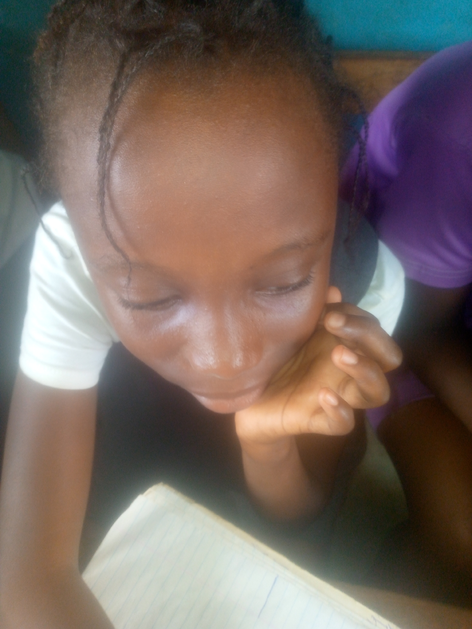 A close-up, high-angle shot shows a young Black girl resting her chin on her right hand. She wears a white t-shirt and is positioned near a notebook with blue lines. Part of a person wearing a purple garment is visible to the right. The location is Unknown, Unknown.