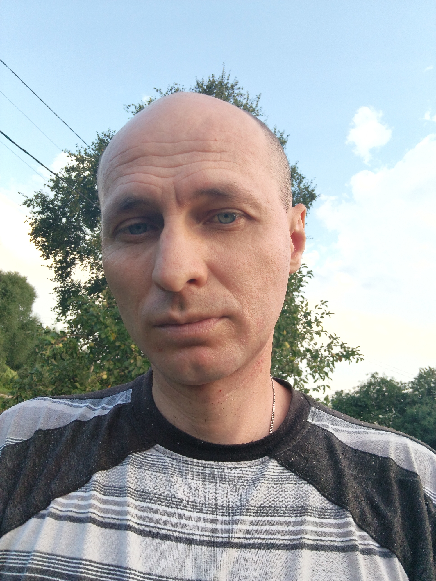 A close-up, head-and-shoulders selfie shows a bald, light-skinned man with blue eyes, a neutral expression, and visible facial pores. He is wearing a horizontally striped t-shirt with grey and white bands, and a thin silver chain around his neck. The background is an outdoor scene with a soft focus on trees and a pale blue sky with scattered white clouds. The image appears to have been taken during daylight hours. The location is identified as Mokshan, Russia.
