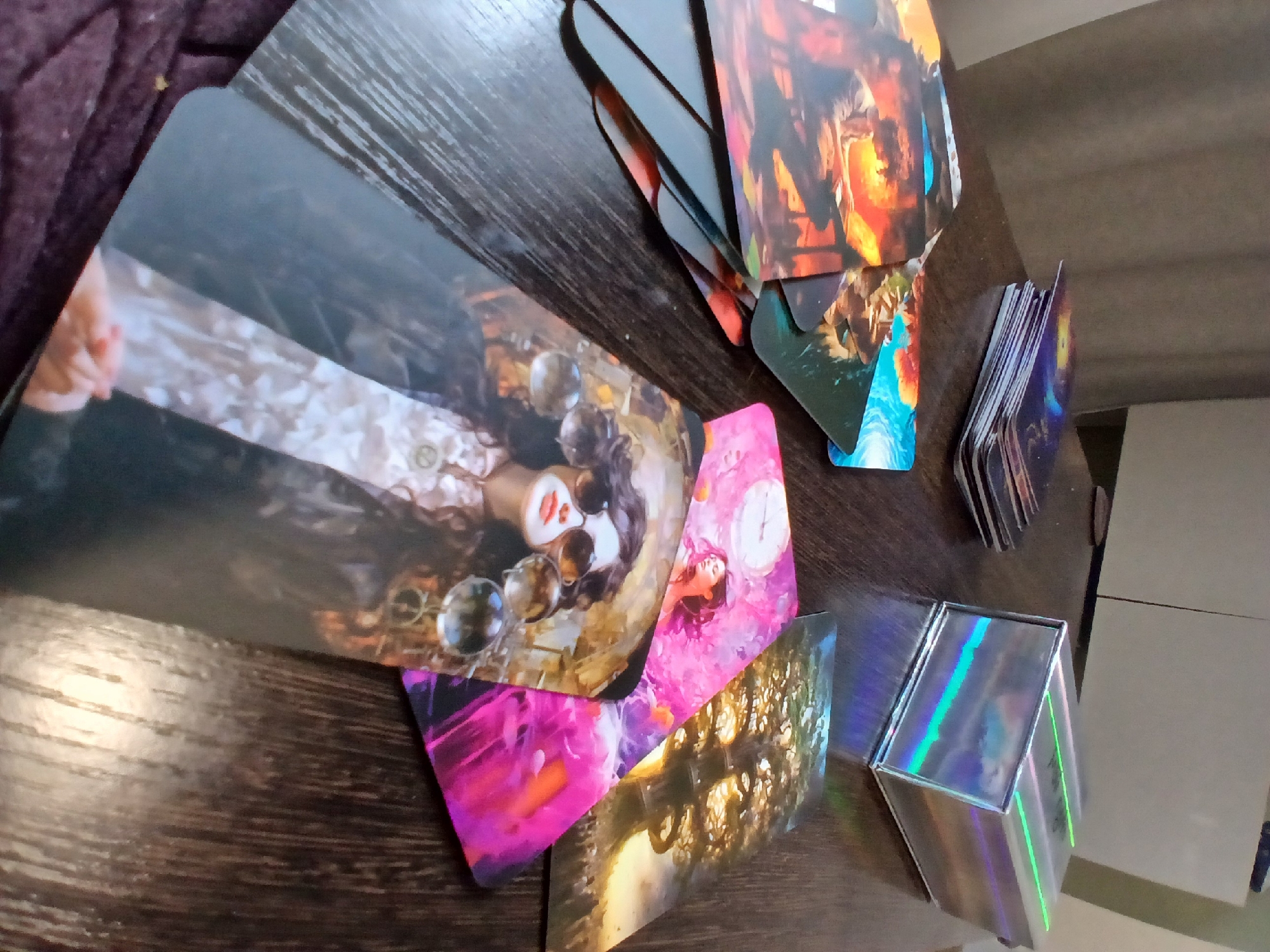 A close-up, angled shot shows a dark wooden table with several tarot cards spread out and a holographic box. In the foreground, a tarot card depicts a woman with dark, curly hair, wearing sunglasses and a patterned shirt, with several translucent orbs in front of her. To the upper left, a stack of tarot cards is partially visible, with a holographic box to the right of them. Several other tarot cards are scattered across the table, displaying abstract and figurative artwork, including one with a large tree. The image has a shallow depth of field, with the focus sharpest on the card in the foreground and the holographic box. The lighting appears to be from an overhead source, casting reflections on the box and cards. There is no visible connection to Yekaterinburg, Russia, or any specific urban environment. The scene is indoors, with a hint of a curtain or fabric in the background.