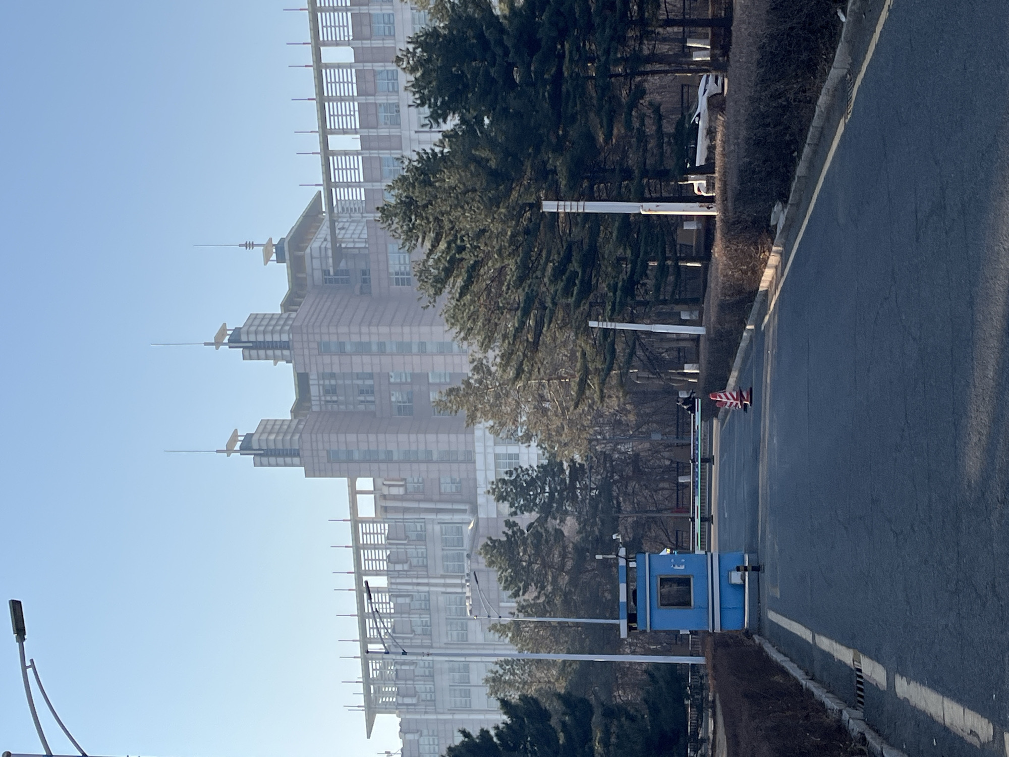 The image shows a street in Changchun City, China. On the left side of the street is a large building with multiple towers and spires. The building appears to be constructed of a light-colored stone or brick. In the foreground, there is a blue guard shack with a window and a barrier gate across the road. The street is paved with asphalt and has a white line marking the edge. There are trees and bushes along both sides of the street. The sky is clear and blue. The image has a slight haze or fog, which may indicate air pollution. There are lampposts lining the street on the left, and a white vehicle is partially visible in the background. A red and white striped traffic cone is placed on the street.