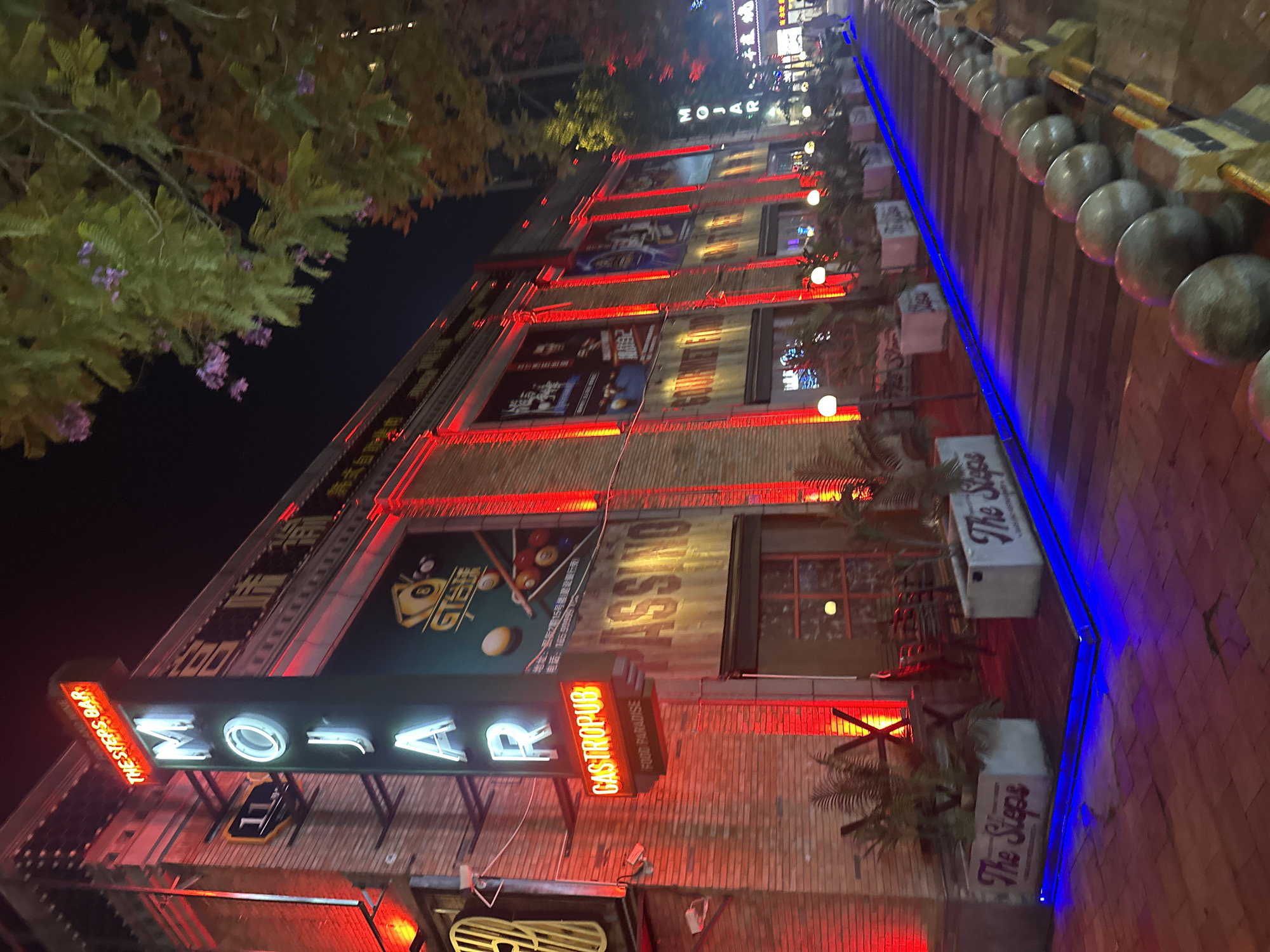 The image shows the exterior of "MOJAR GASTROPUB" in Chongqing High-tech Zone, China, at night. The building is brick and has multiple stories. Red lights outline each story. Signage includes "MOJAR," "THE STEPS BAR," "GASTROPUB," and "FOOD PARADISE." There are advertisements on the building's facade. Planters labeled "The Steps" are visible near the sidewalk. The sidewalk is brick and has blue lighting along the edge. There are spherical concrete barriers along the sidewalk. Trees with flowers are visible on the left side of the frame. The overall scene appears to be a lively nighttime view of the bar's location.