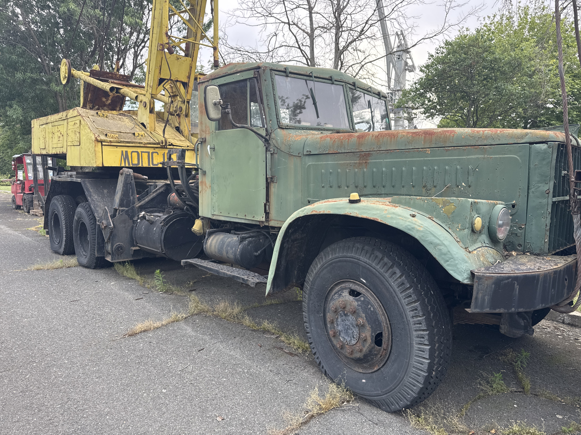 This media file showcases a weathered, vintage green crane truck parked on an asphalt surface. The truck, which appears to be in a state of disrepair, is the primary object in focus, with its front portion and part of its crane clearly visible. The crane is yellow and also shows signs of age and rust. In the background, there are trees with bare branches, suggesting a possibly cool or overcast day, and a hint of industrial or industrial-adjacent scenery with other large structures visible.
Notable details include the peeling green paint on the truck's body, the prominent rust patches, and the large, rugged tires. The truck has a distinct, old-fashioned design. On the yellow crane, the word "MONOLIT" is visible in black letters, indicating the name of a company or brand. The scene is outdoors, and the lighting suggests it might be daytime, but the overcast sky contributes to a somewhat somber mood, emphasizing the neglected state of the machinery. The location is identified as Burgas, Bulgaria, though no specific landmarks are immediately identifiable within the frame to confirm this. There are no people visible in the image, and no specific activity is taking place; the truck is simply parked.