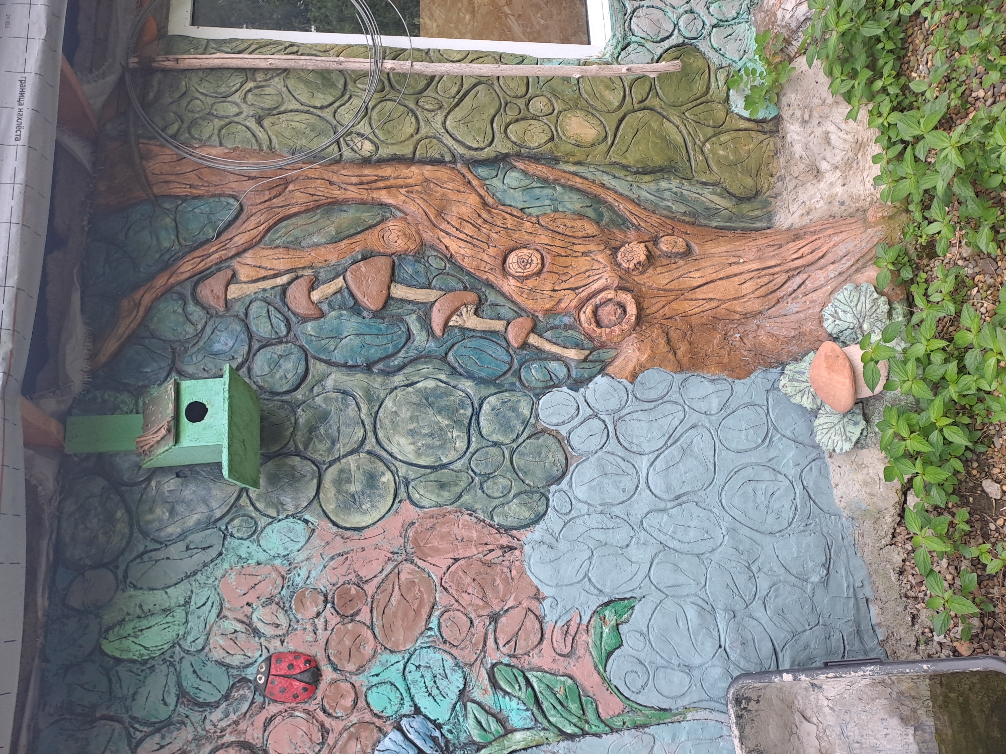 The image shows a section of an exterior wall decorated with a painted mural of nature. The mural features a ladybug, a birdhouse, leaves, mushrooms, and a tree trunk. The wall itself is textured, with raised circular and leaf-like patterns. The area at the bottom of the image shows gravel and green plants. A wheelbarrow is partially visible on the left. A window is visible on the right side of the image.
