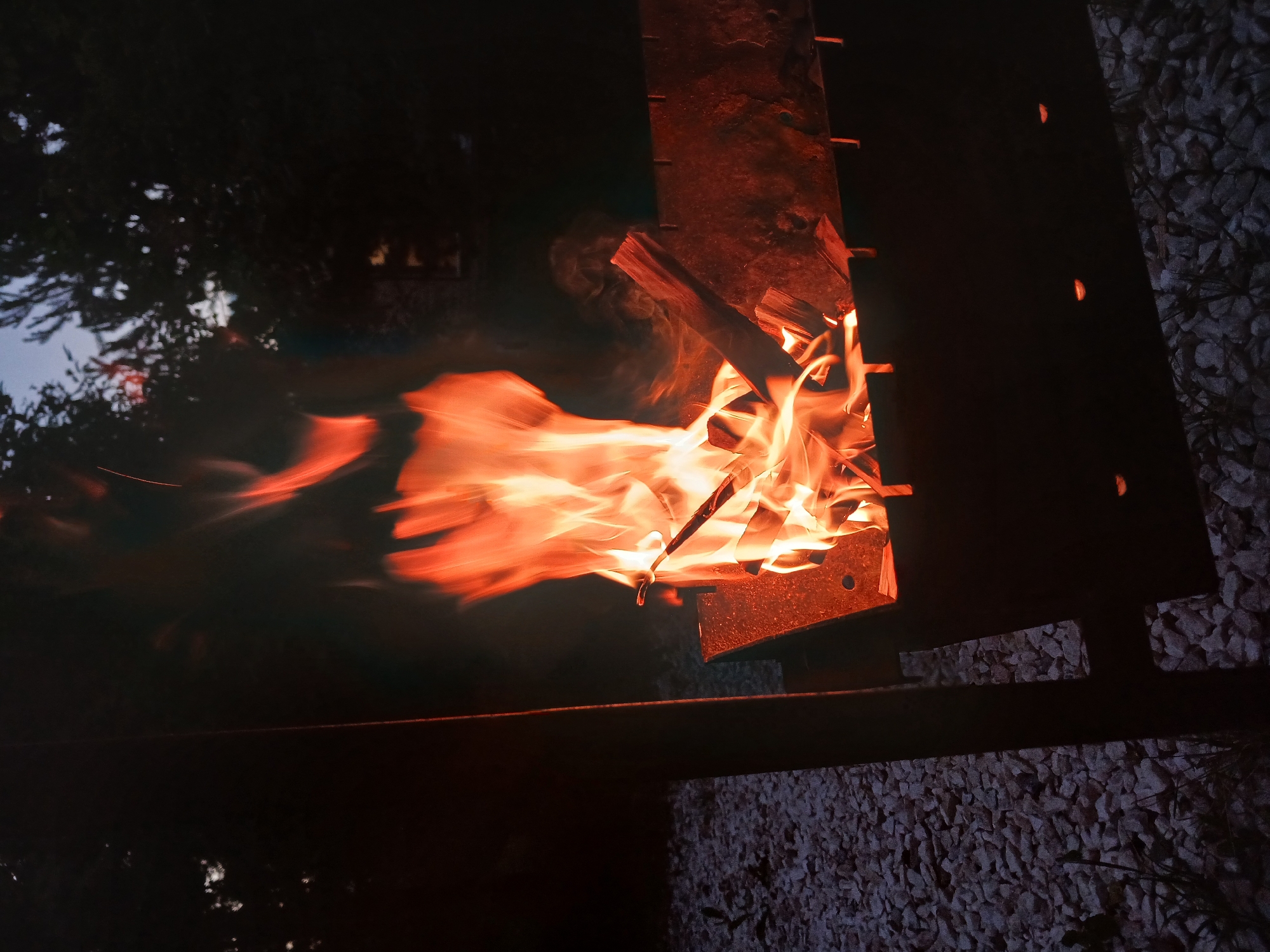 The image shows a close-up view of a fire burning in what appears to be an outdoor fire pit or barbecue grill. The fire is vibrant orange and yellow, with wisps of smoke curling upwards. Several pieces of wood are visible, some partially consumed by the flames. The fire pit itself is constructed from dark metal, with a rough, textured surface and visible rivets or bolts along its edge.
In the background, to the left of the fire, a dark, blurry area suggests foliage or trees, possibly illuminated by the fire's glow. The sky appears dark, indicating it is either dusk or nighttime. To the right and bottom of the frame, a textured surface of small stones or gravel is visible, further suggesting an outdoor setting. The overall mood is warm and perhaps a bit wild, given the dynamic nature of the flames. There is no visible text in the image. The context of Verkhneye Dubrovo, Russia, places this scene in a rural or suburban outdoor environment. The time of day is clearly evening or night due to the darkness and the focus on the firelight.