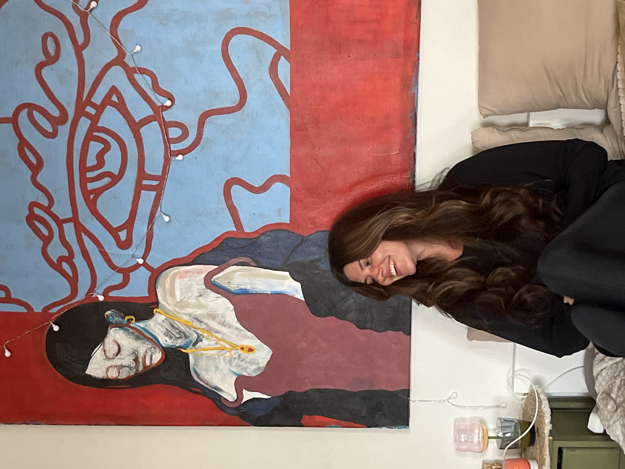 A woman with long brown hair is smiling and leaning toward an abstract painting. The painting is divided into a blue and red background. The blue section has red abstract lines resembling an eye and other shapes, and the red section features a person lying down. The woman is wearing black clothing and appears to be indoors. There are string lights attached to the painting. A small decorative lamp and other objects are visible at the bottom of the image. The location is unknown and cannot be determined from the image. The city of Krasnoyarsk, Russia, is not discernable from the details in the scene.