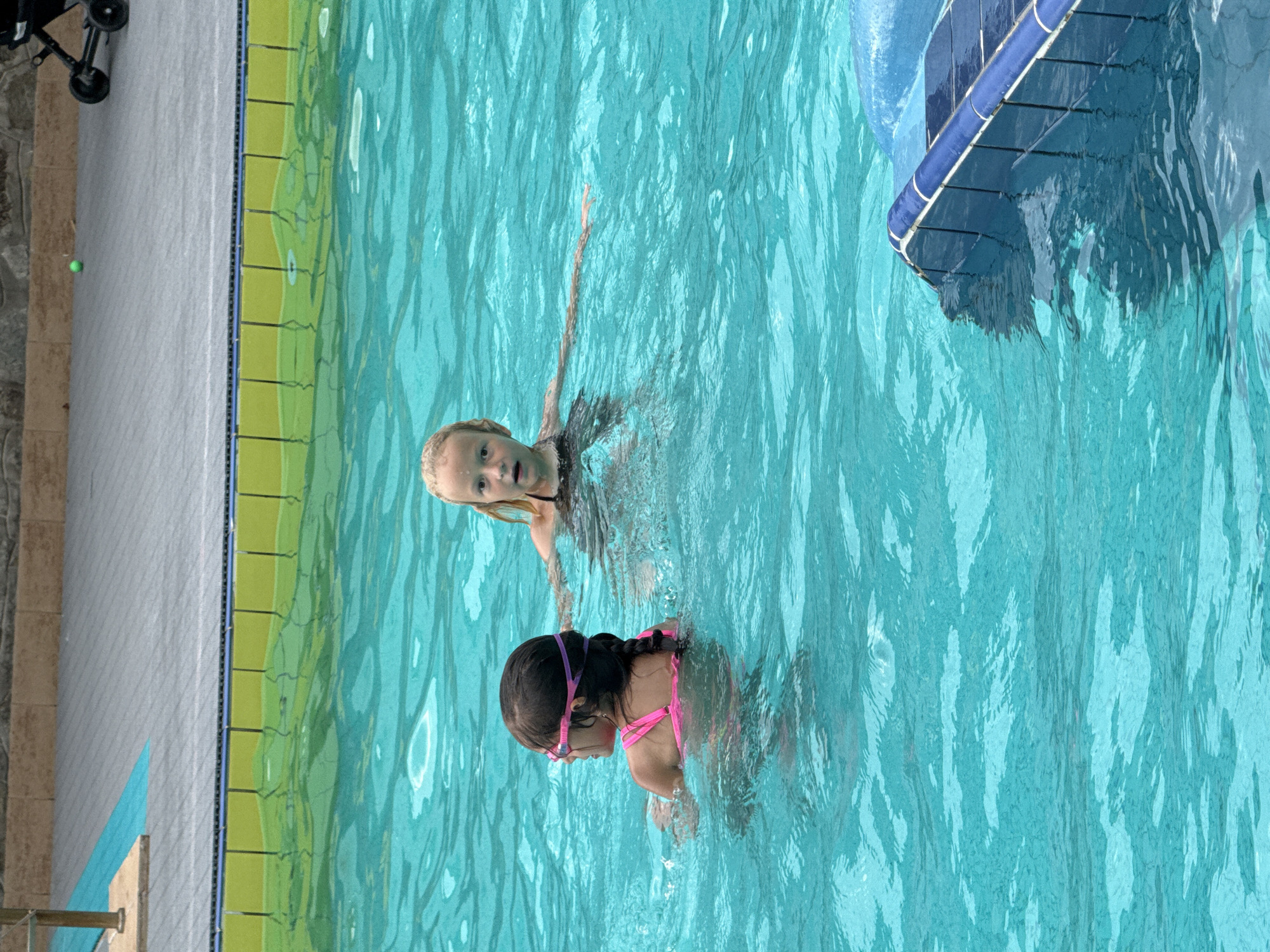 Two young girls are swimming in a pool. The girl in the foreground on the left is wearing a pink swimsuit and goggles, and has her hair in a braid. The girl on the right has blonde hair and is wearing a dark swimsuit. The pool water is a bright turquoise. In the background, there is a paved area with a stone wall and a stroller. The location is likely an outdoor swimming pool.
