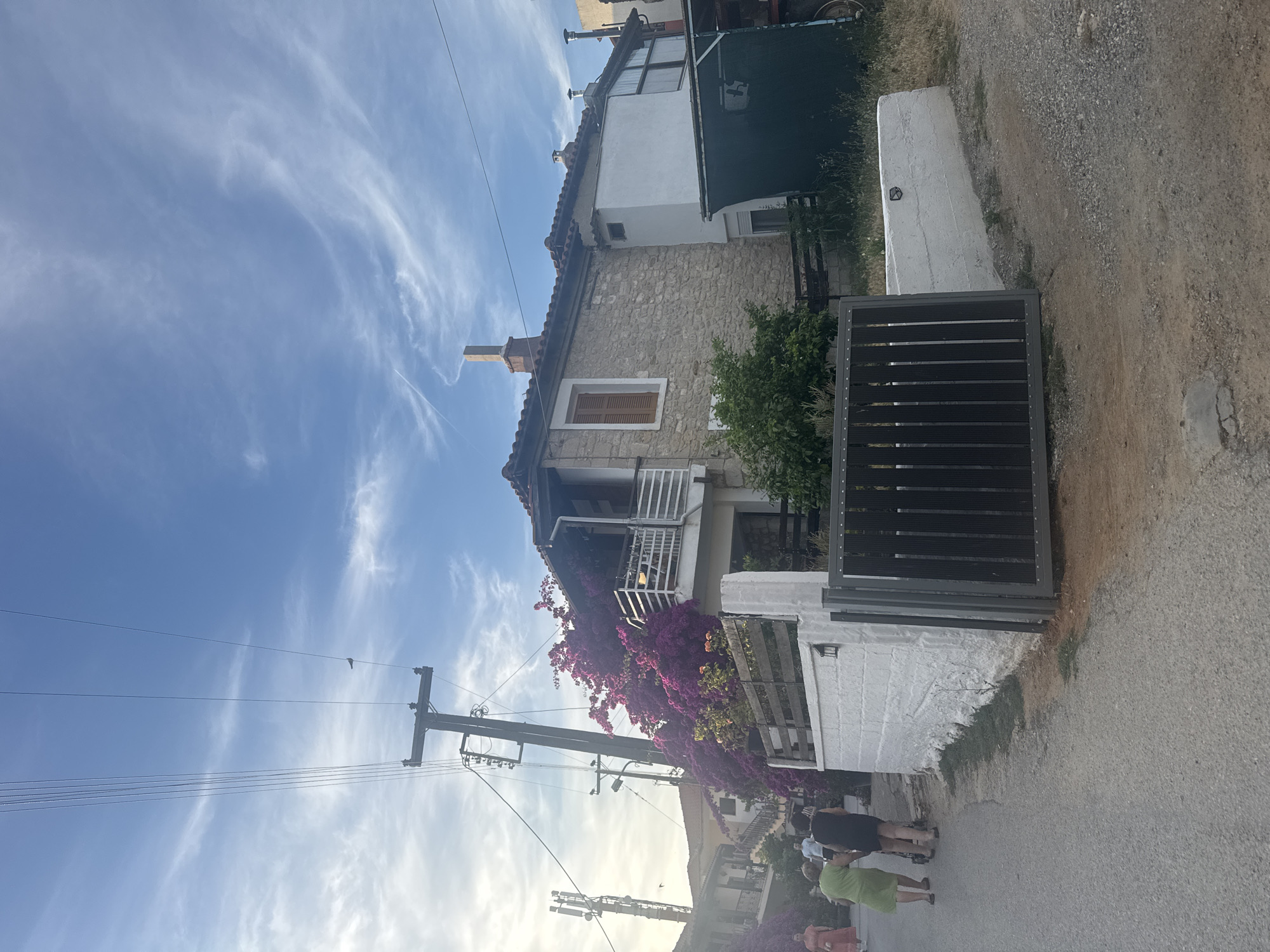 In the city of Kassandra Municipal Unit, Greece, a village road curves gently to the left, lined with asphalt and gravel. The day is bright, with a clear blue sky scattered with wispy white clouds. A prominent wooden utility pole, adorned with insulators and wires, stands on the left side of the road, alongside a modern cell tower.
Several people are visible on the road. Two women, one in a lime green dress and the other in a black dress, walk away from the camera. A third woman in a pink dress is further down the road. In the background, a traditional stone house with a red-tiled roof and a white-railed balcony is partially obscured by vibrant purple bougainvillea. Adjacent to it, a white building stands next to a dark green tarp covering an unseen object. A dark metal gate guards the entrance to the property. The scene is characterized by its rustic charm and a blend of natural beauty and everyday village life.